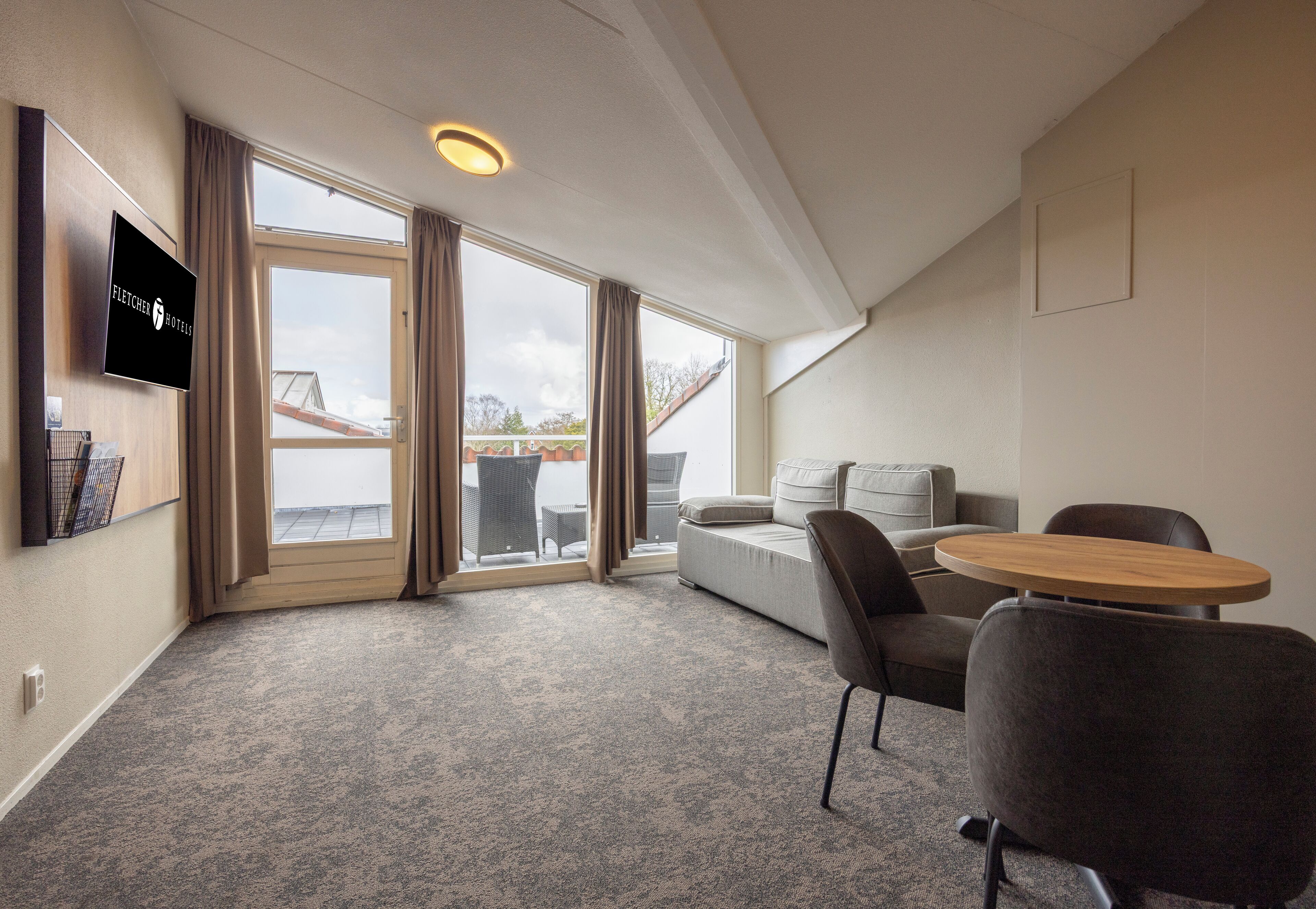 A modern hotel room with a large window, a flat-screen TV, a sofa, a round table, and a carpeted floor.