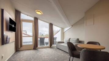 A modern hotel room with a large window, a flat-screen TV, a sofa, a round table, and a carpeted floor.