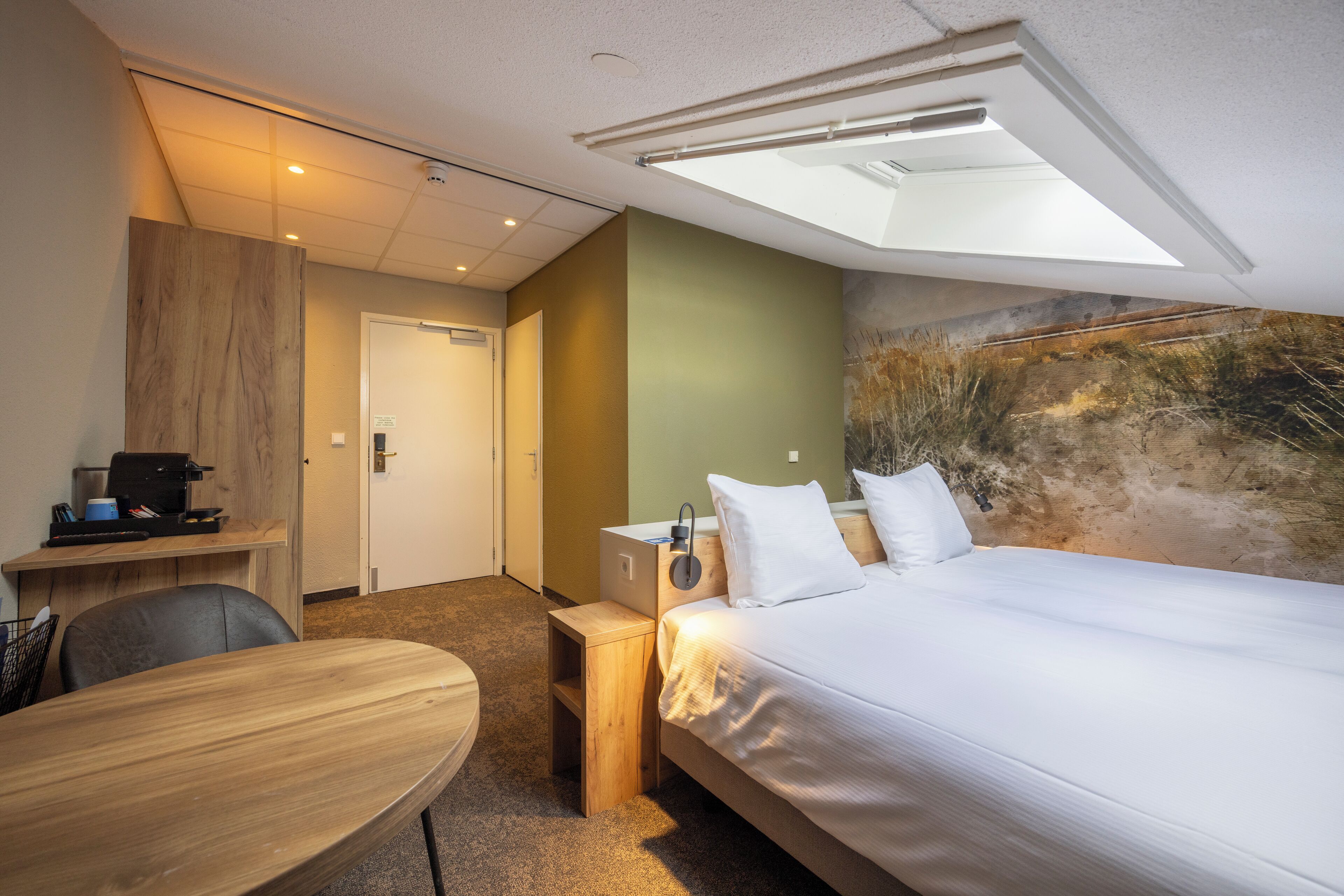 Habitación de hotel con cama, escritorio, silla, mesita pequeña, mesita de noche, lámpara y un cuadro en la pared.