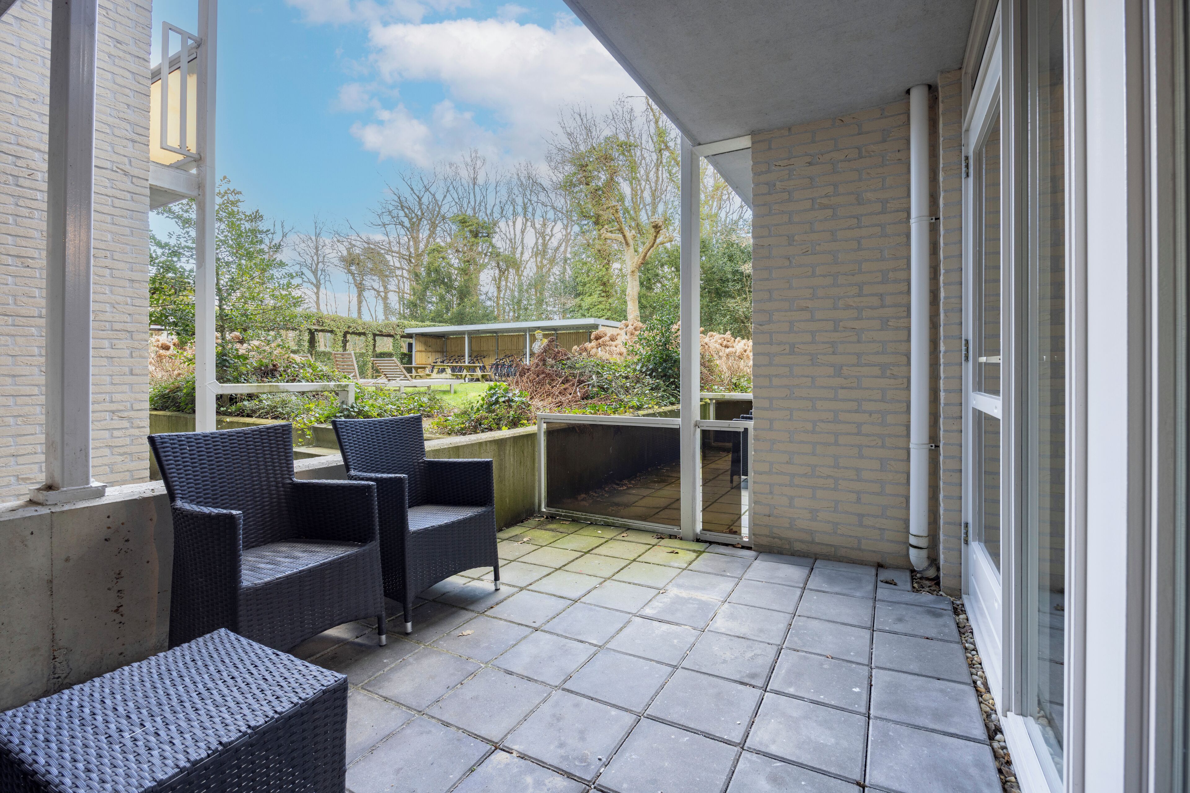 A covered patio with two wicker chairs and a small table, a brick wall, and a view of a garden with a pergola and trees.