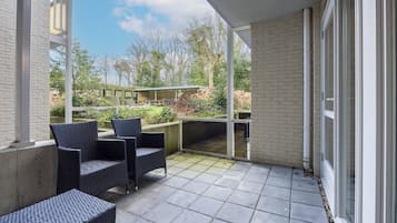 Eine überdachte Terrasse mit zwei Korbstühlen und einem kleinen Tisch, einer Ziegelwand und Blick auf einen Garten mit einer Pergola und Bäumen.