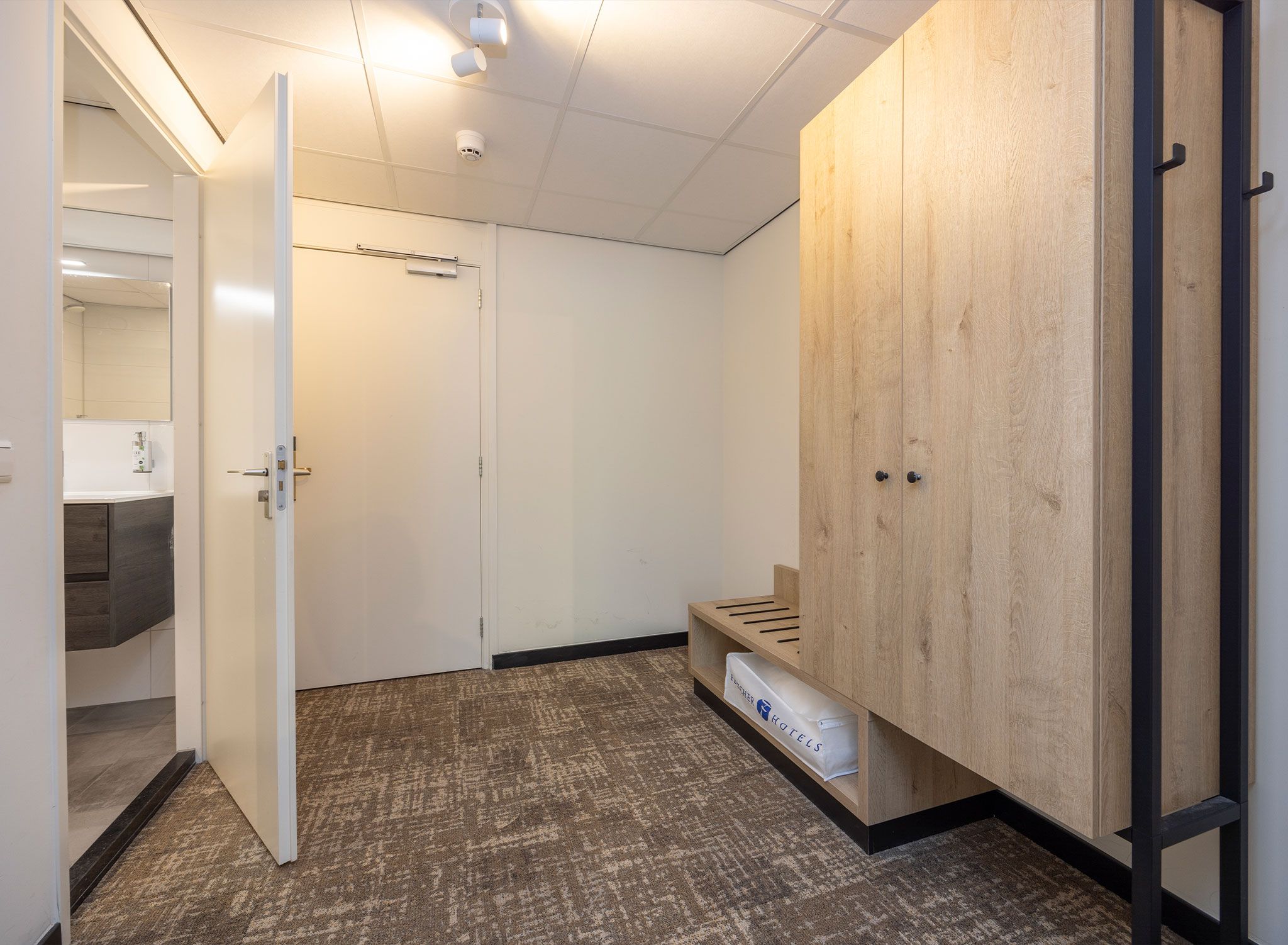 A hotel room with a wooden wardrobe, a bench, and a carpeted floor.