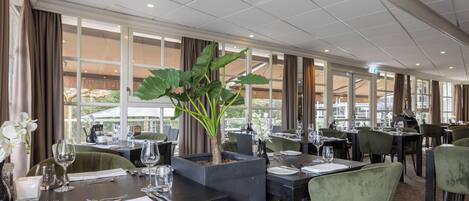 A restaurant with tables set for dining, large windows, and a potted plant.
