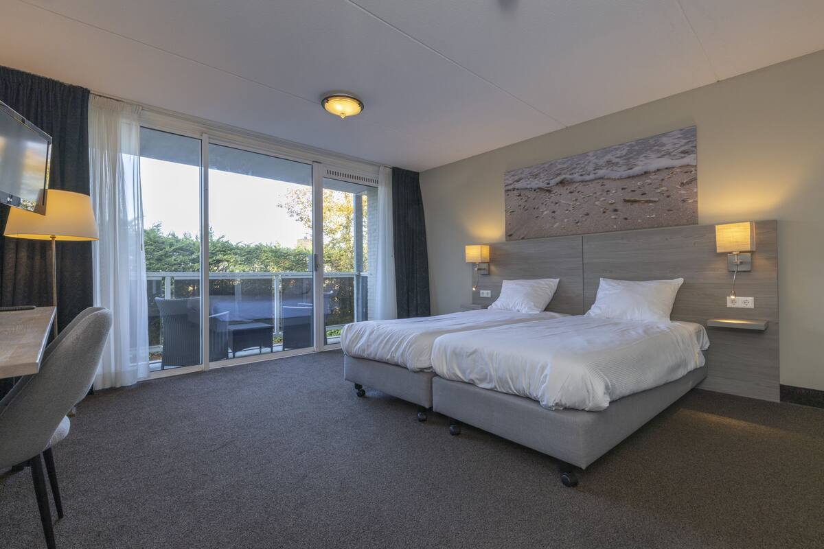 A hotel room with a large bed, a desk, a chair, a balcony with a view, and a wall-mounted artwork.