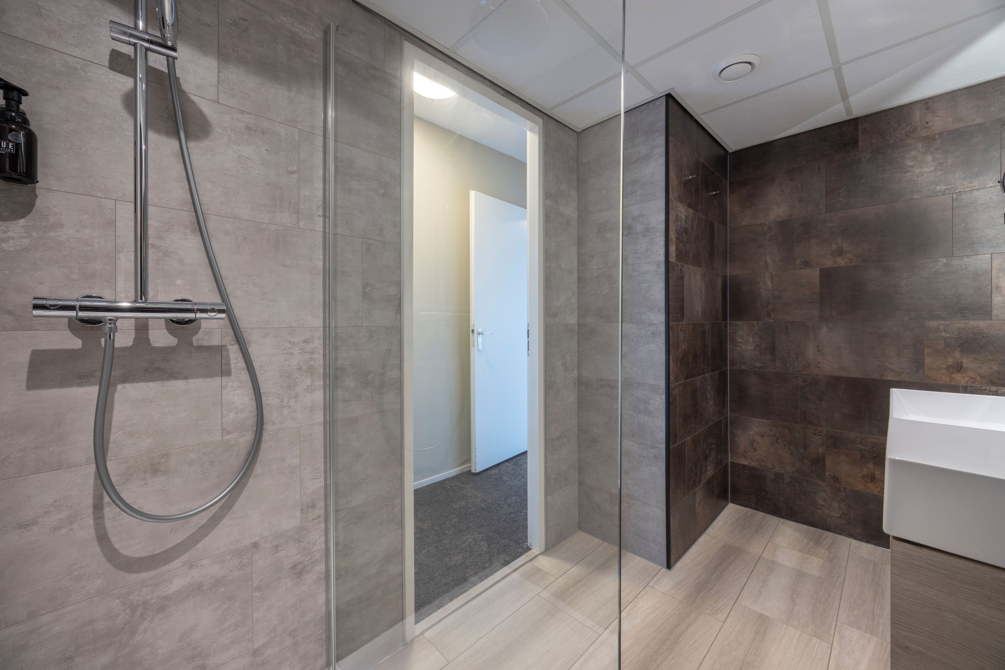 Un baño moderno con cabina de ducha de vidrio, lavabo blanco y paredes cubiertas de baldosas oscuras.