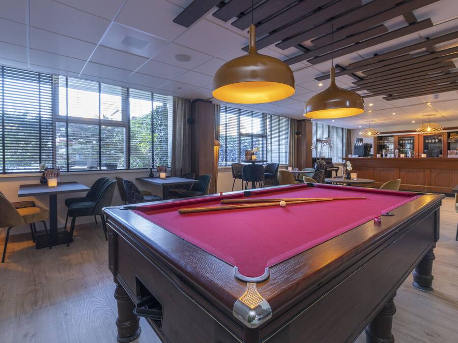 Una mesa de billar con superficie de fieltro rojo, mobiliario de madera e iluminación colgante en una sala con ventanas amplias y una zona de bar.
