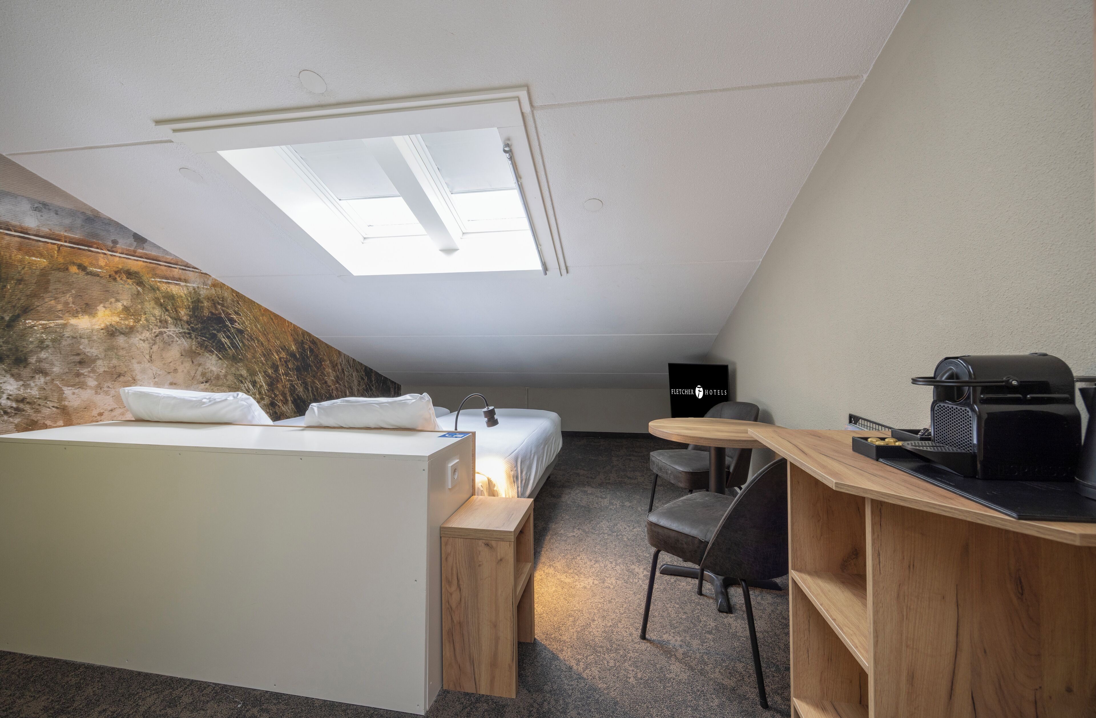 Een kleine hotelkamer met een bed, een bureau, een stoel, een koffieapparaat en een dakraam.