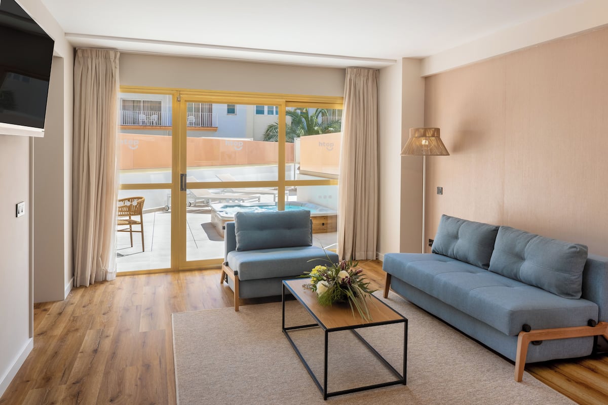 Un salón moderno con sofá, sillón y mesa de centro. Un ventanal corredero da a un espacio exterior con piscina y sillas de descanso.