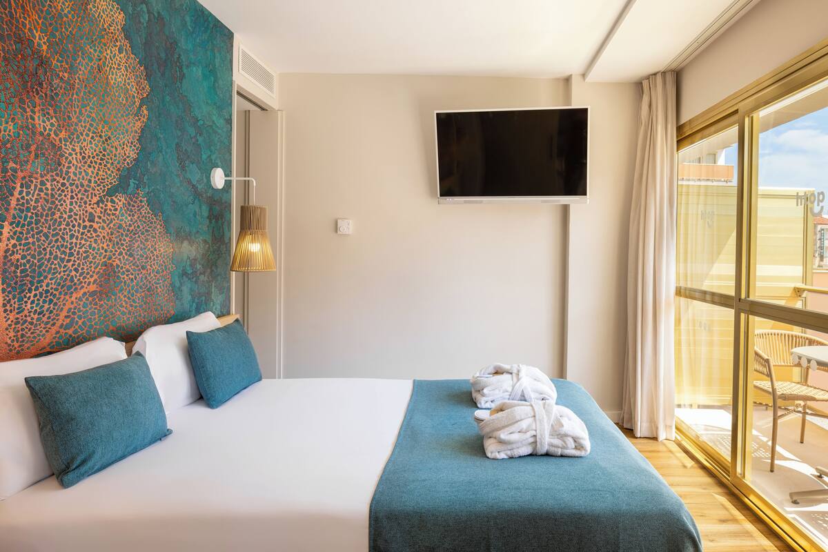 A hotel room with a bed, a TV mounted on the wall, and a view of the outside through large windows.