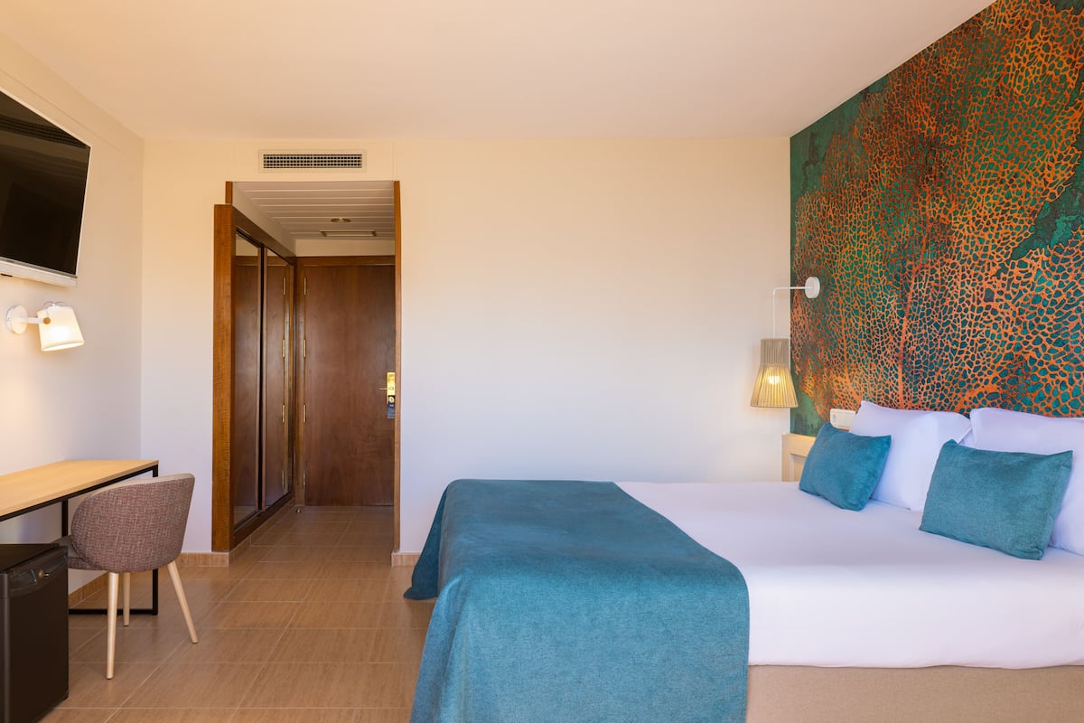 Habitación de hotel con cama, escritorio y silla. Hay un televisor en la pared y un adorno mural con estampado.