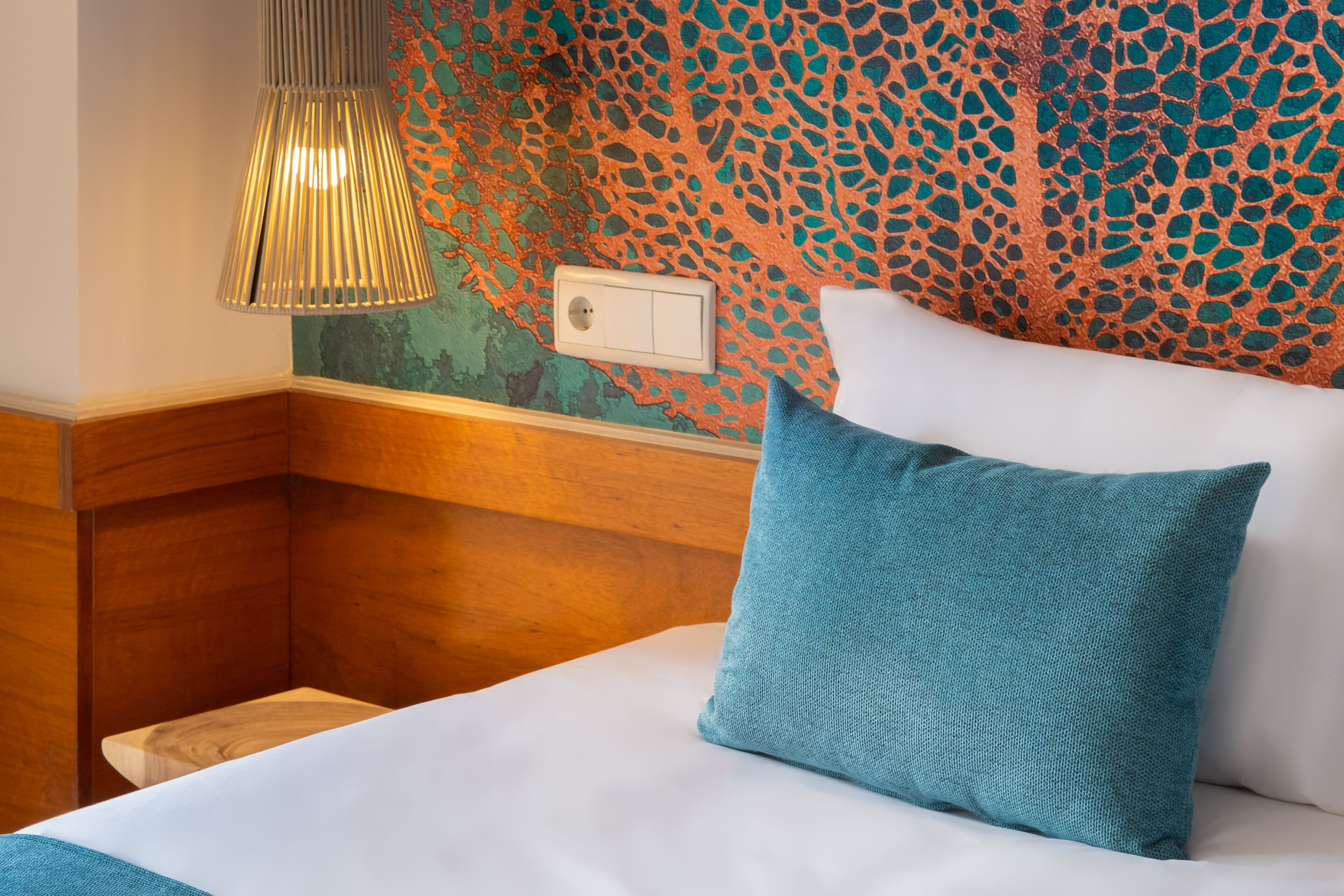 A neatly made bed with white linens and a teal pillow, a textured wall with a mosaic pattern, and a wooden headboard.