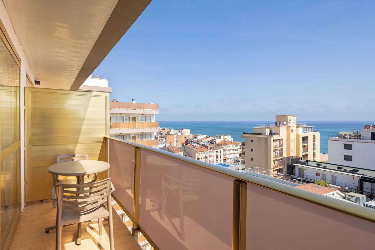 Un balcón con mesa y sillas, con vistas a una ciudad costera con edificios y el mar.