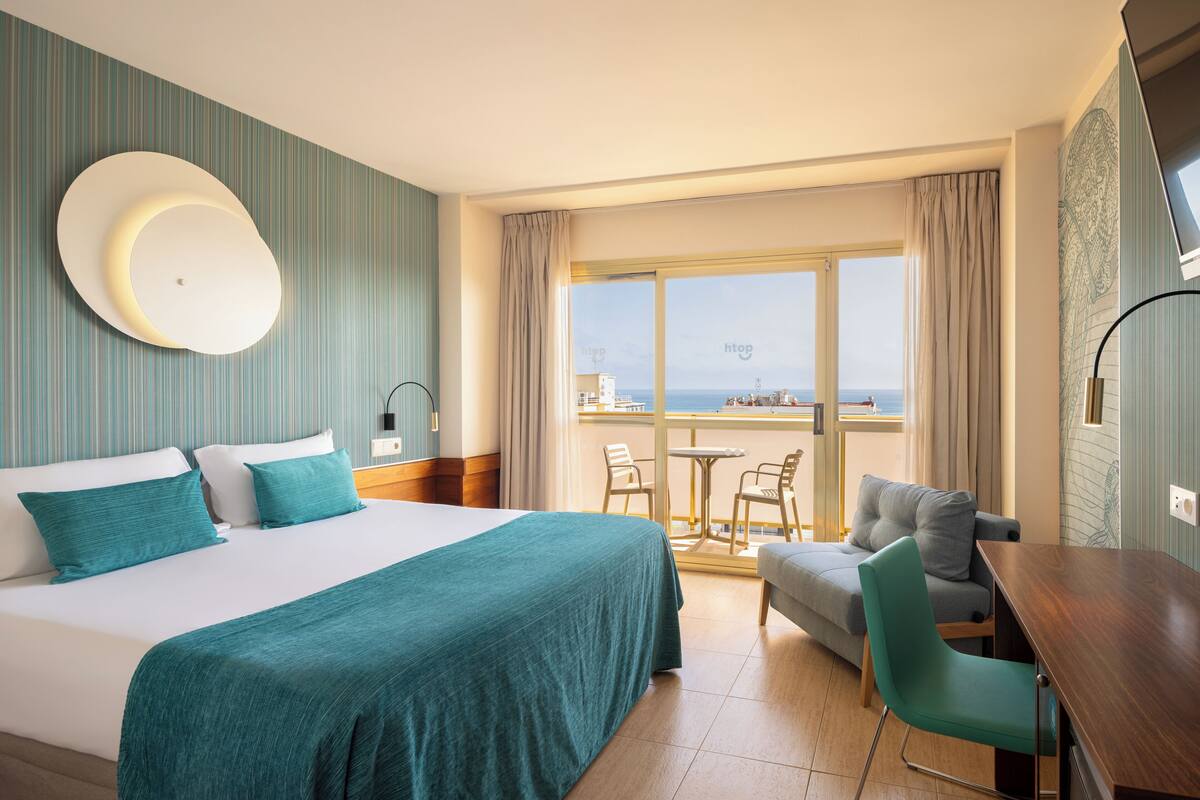 Habitación de hotel con una cama grande, un sofá, una mesa de comedor y un balcón con vistas al mar.