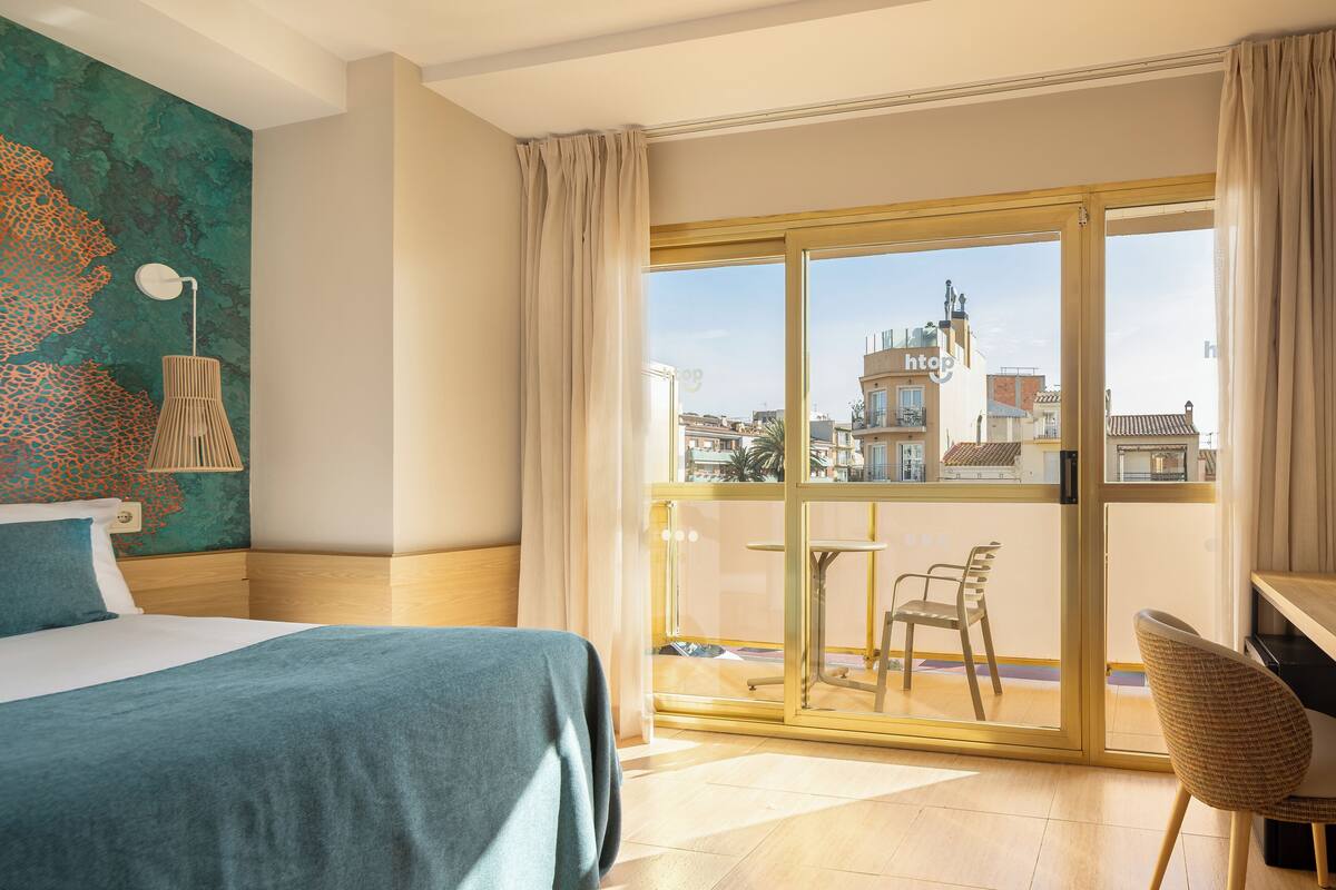 Habitación de hotel con cama, escritorio, silla, balcón con mesa y sillas, y vista a edificios.