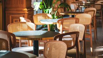 A restaurant interior with round tables and wicker chairs, wooden paneling, and green plants.