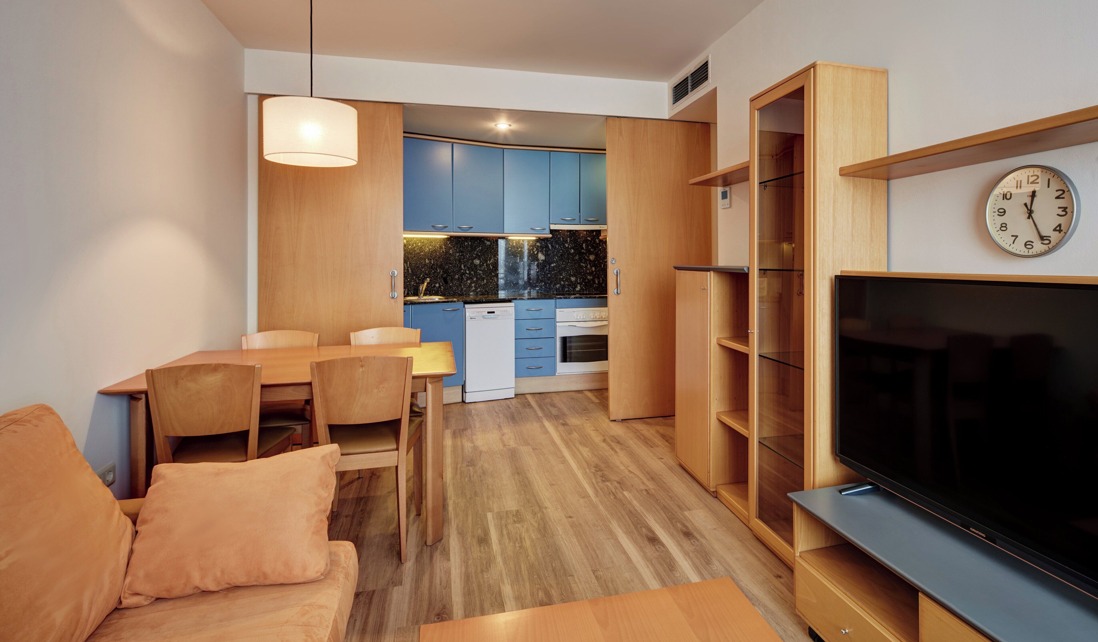 A modern living room with a flat-screen TV, wooden furniture, and a dining area with a kitchen in the background.