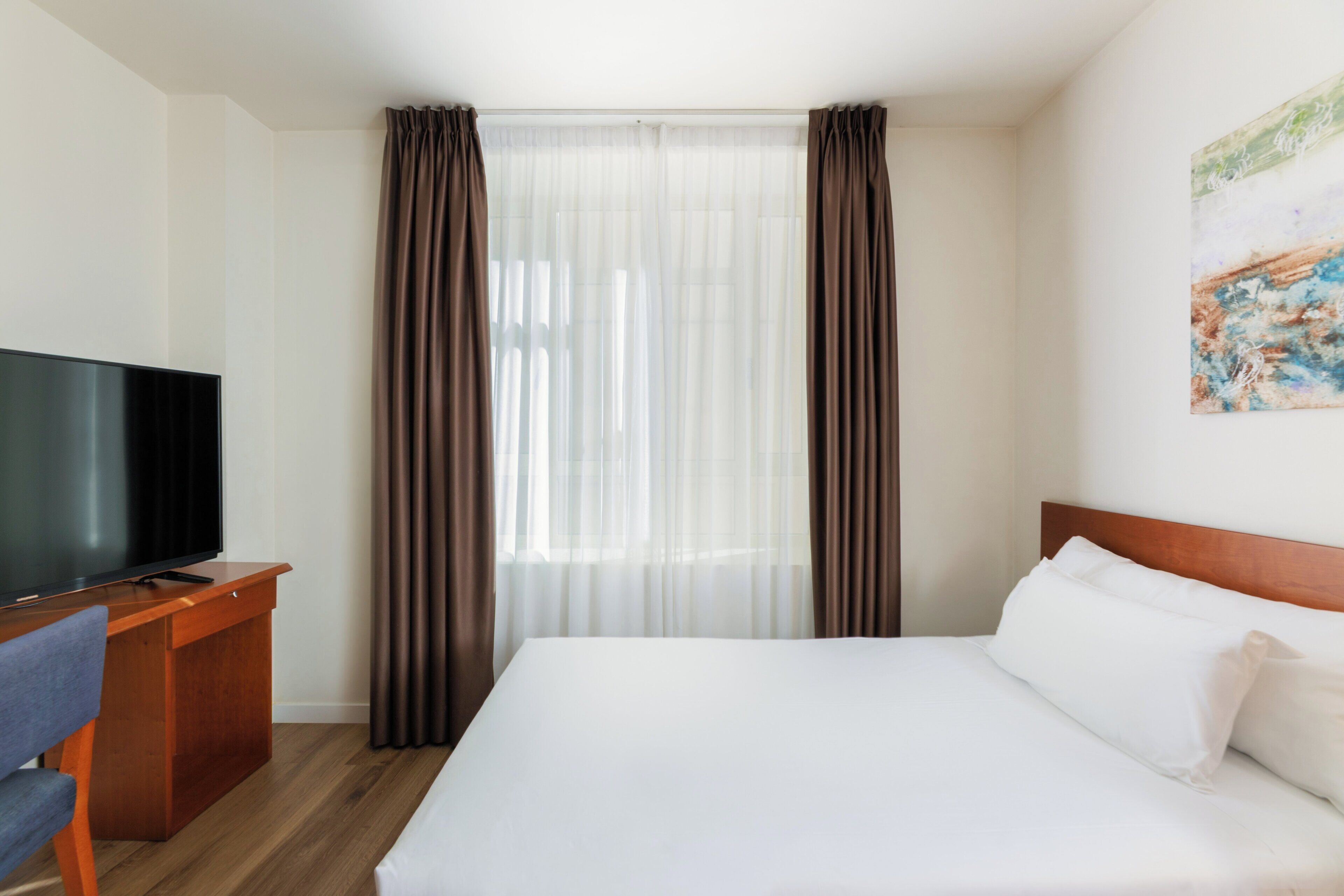 A hotel room with a bed, a TV on a wooden desk, and curtains.
