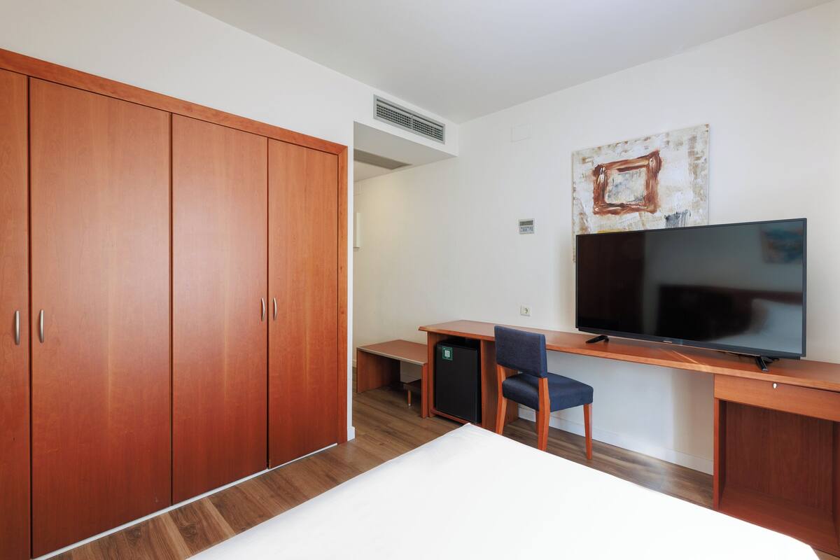 A hotel room with a bed, a desk with a chair, a television, and wooden wardrobes.