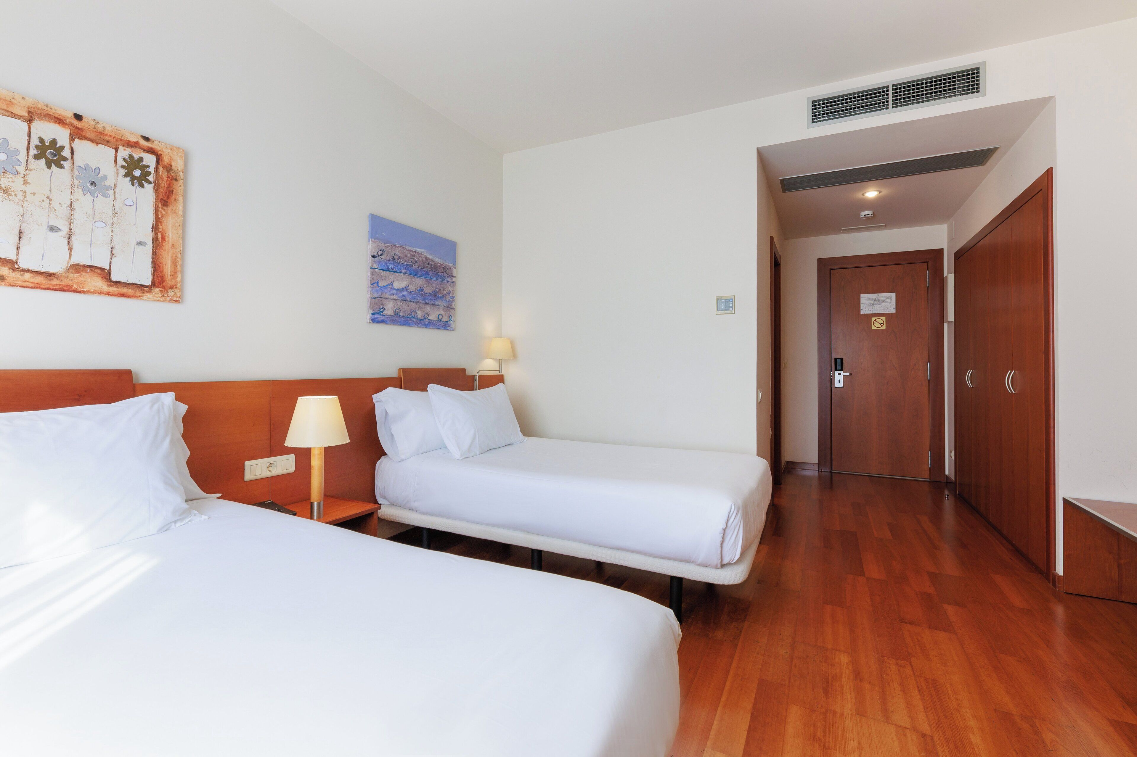 Habitación de hotel con dos camas, cabecero de madera, mesita de noche con lámpara, un cuadro en la pared y un armario de madera.