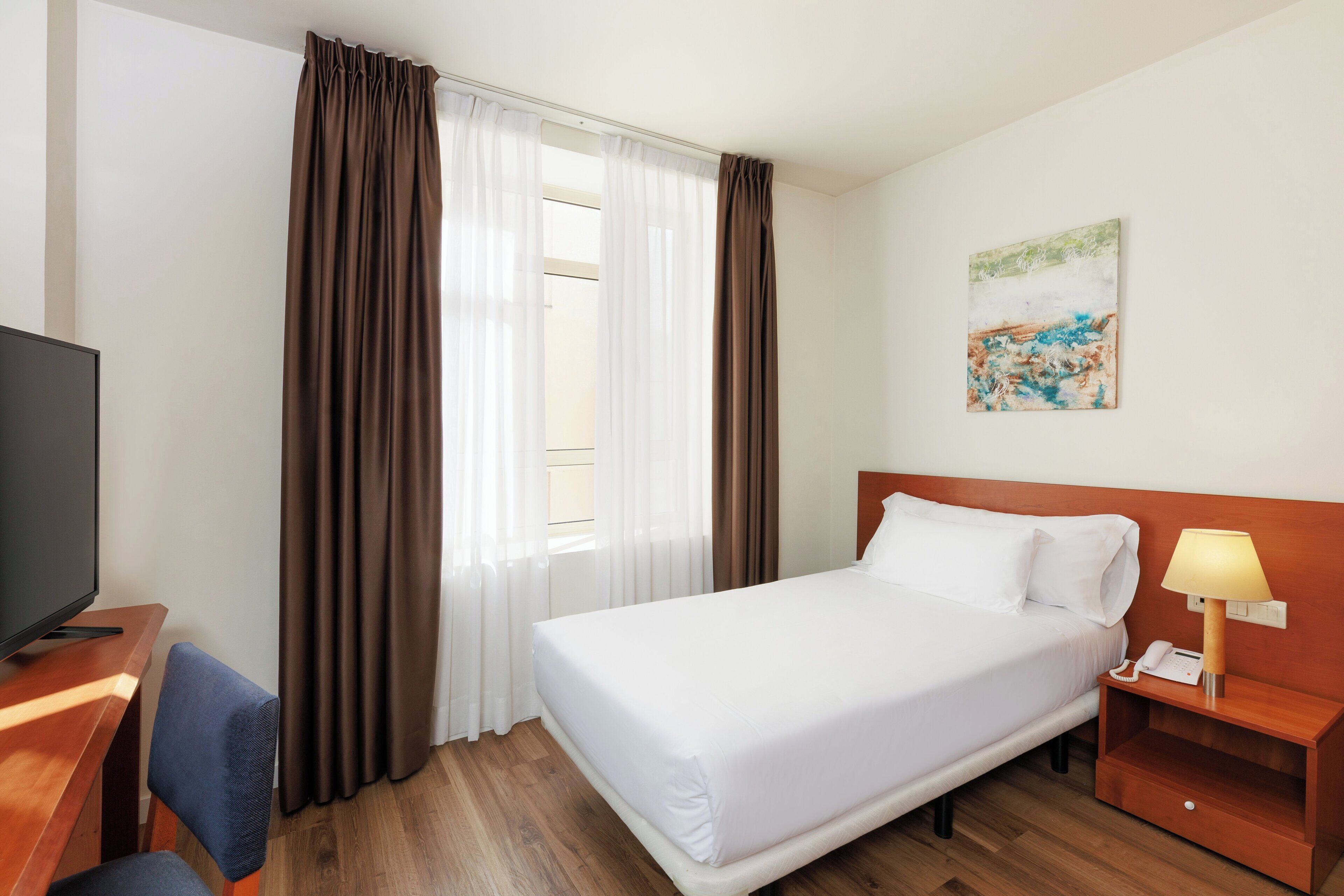 A hotel room with a bed, a desk with a television, a chair, a nightstand with a lamp, and a painting on the wall.