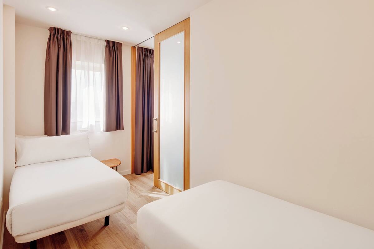 A hotel room with two single beds, a wooden floor, a small bedside table, and a sliding door.
