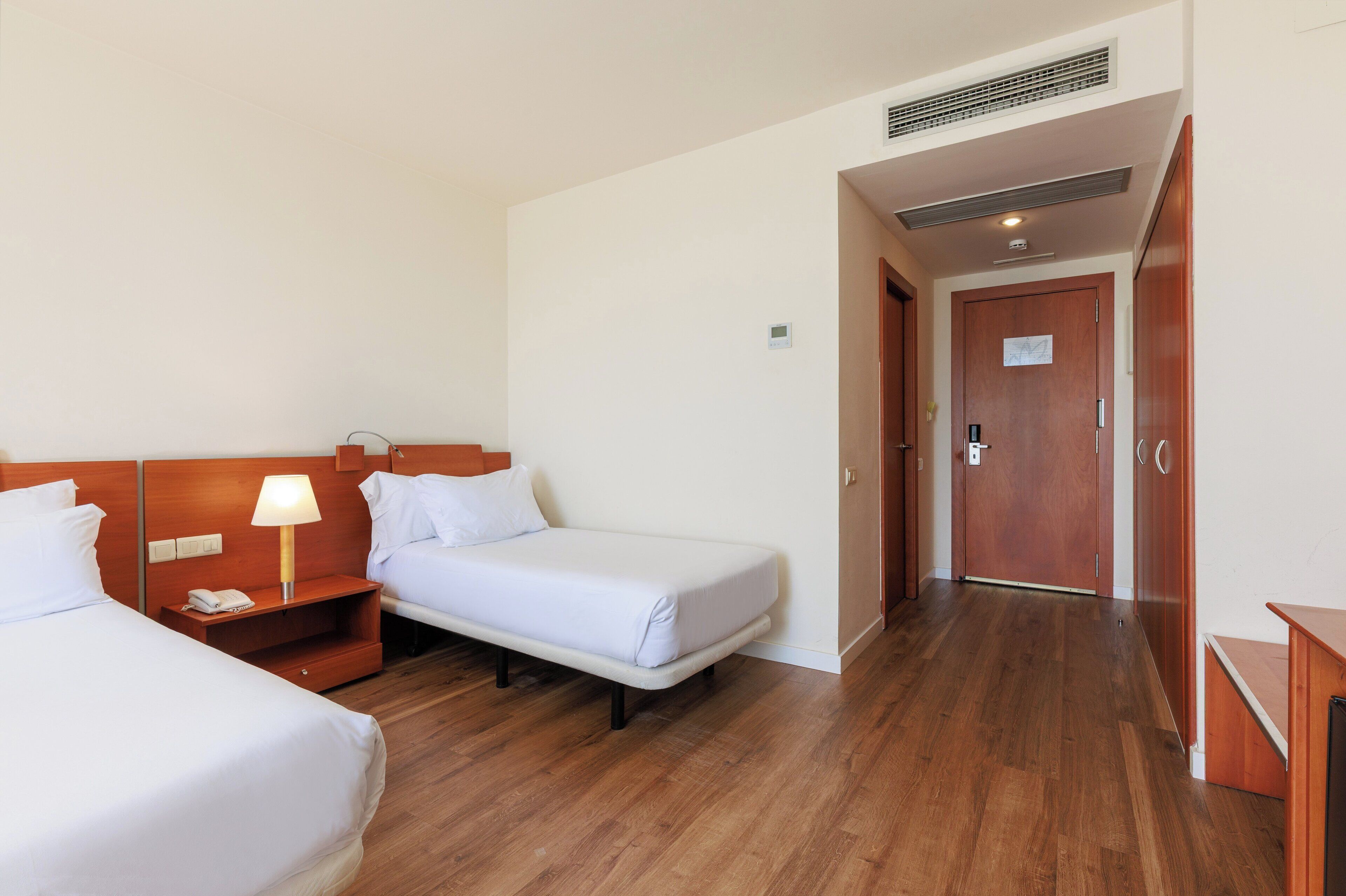 Une chambre d’hôtel avec deux lits simples, une table de chevet avec une lampe, un plancher en bois et une porte donnant sur une autre pièce.
