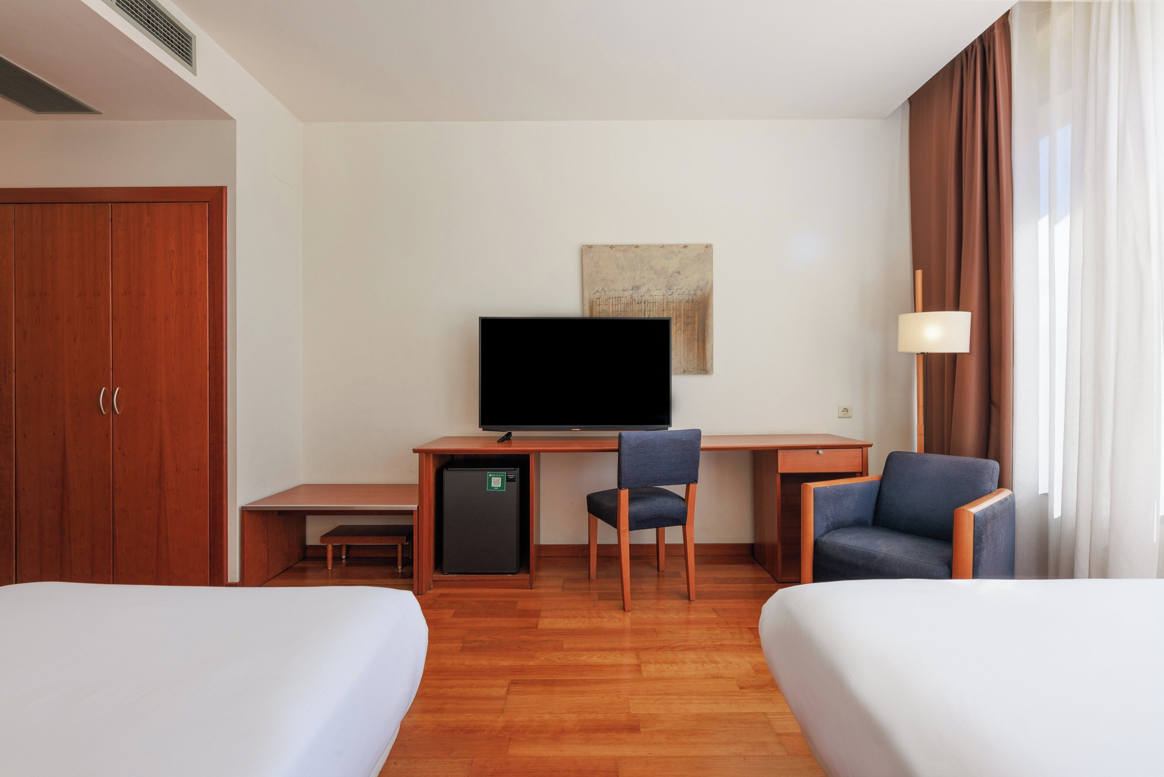 Quarto de hotel com duas camas, uma escrivaninha, televisão, uma cadeira, frigorífico pequeno e uma luminária.