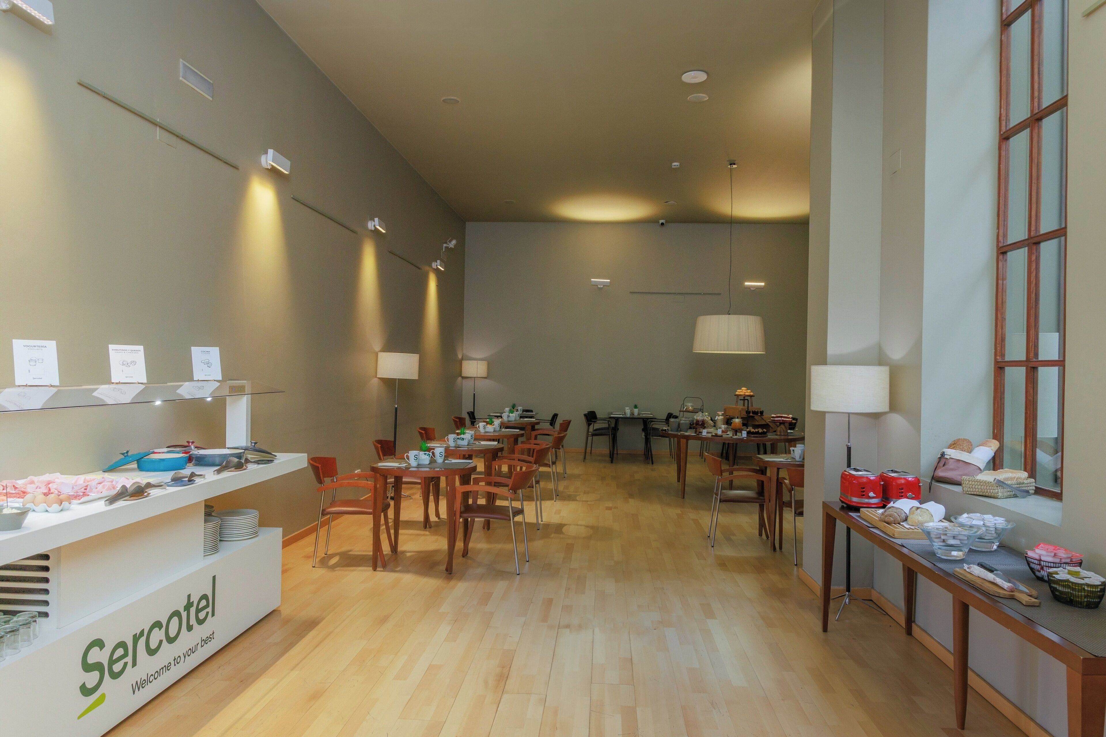 Een ruime eetruimte met meerdere tafels en stoelen, een receptiebalie met de tekst 'Sercotel' en een groot raam aan de rechterkant.