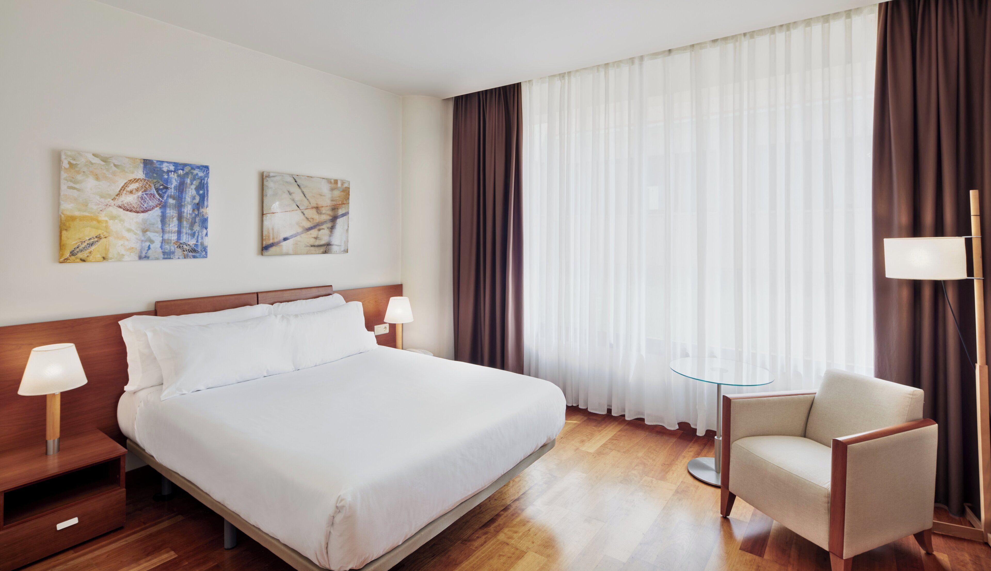 A hotel room with a large bed, two bedside lamps, a chair, a small table, and two pieces of wall art.