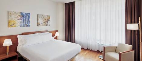 A hotel room with a large bed, two bedside lamps, a chair, a small table, and two pieces of wall art.