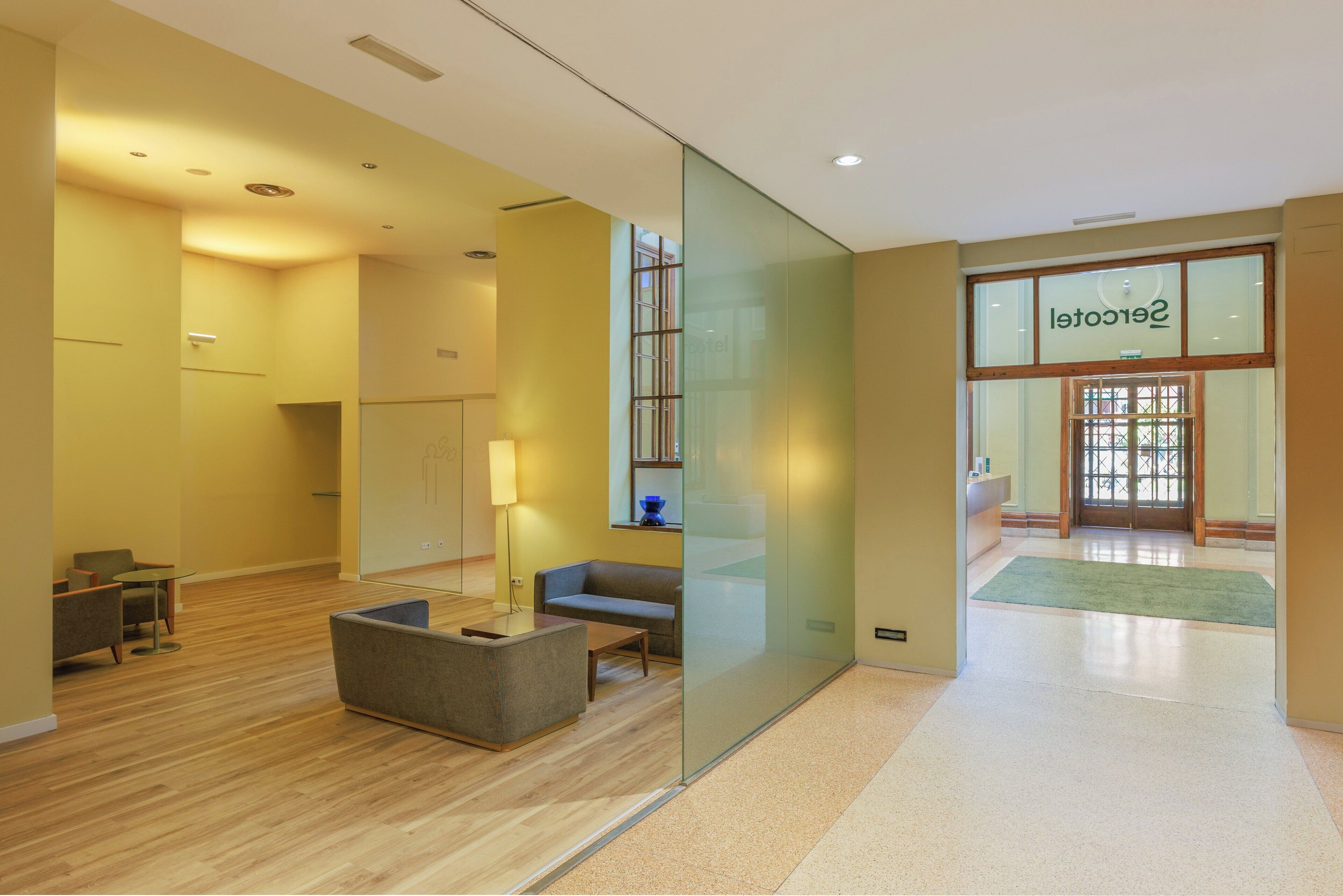 Un moderno hall de hotel con piso de madera, un área de estar con sofá y sillones, una partición de vidrio y un mostrador de recepción.