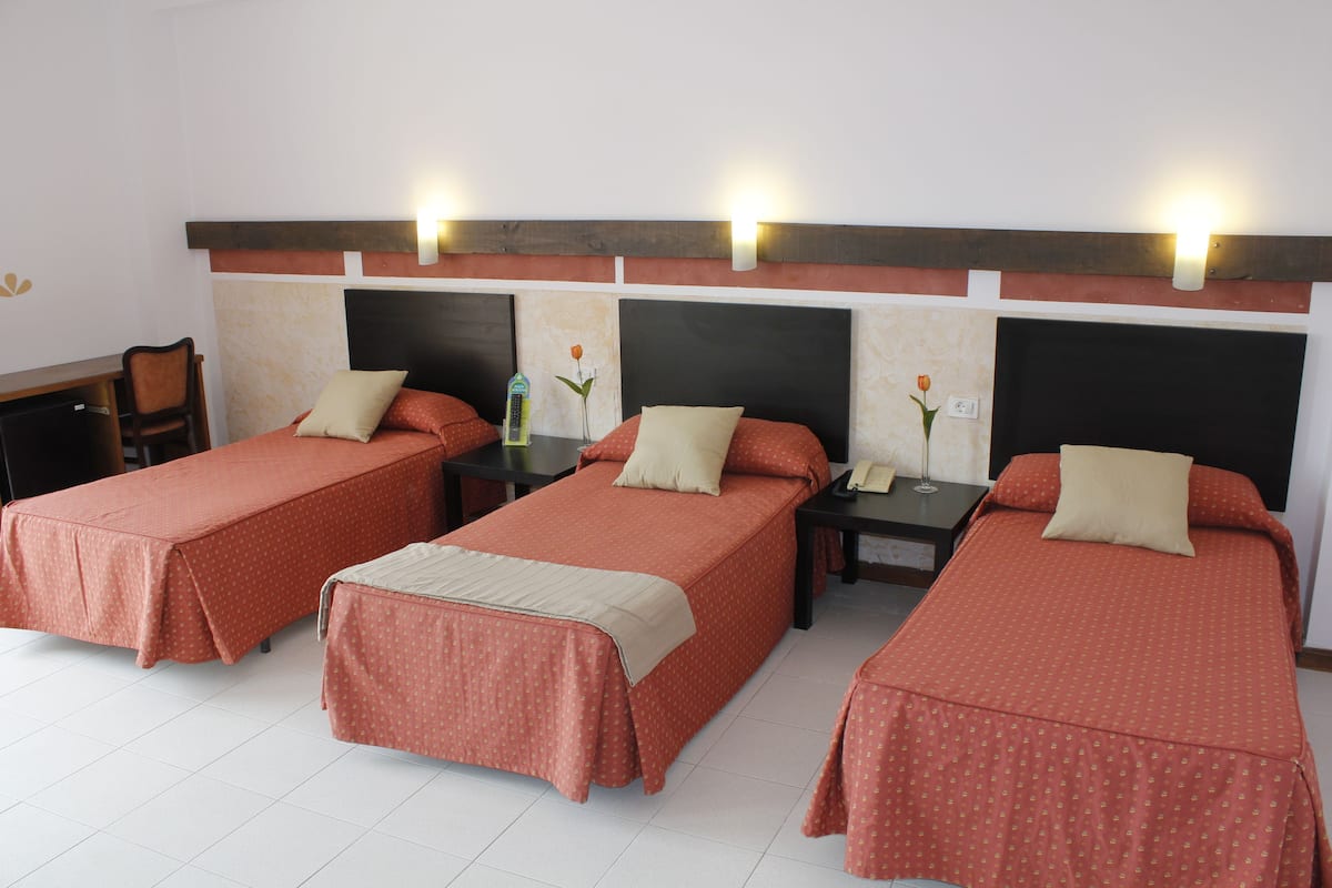 Une chambre d’hôtel avec trois lits simples, chacun recouvert d’une couverture rouge et agrémenté d’un oreiller beige. On y trouve des tables de chevet avec des lampes, ainsi qu’une petite table sur laquelle est posé un vase de fleurs.