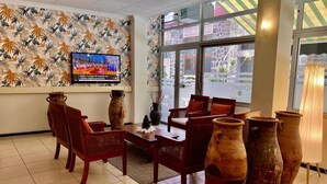 Een lobby met een televisie waarop een nieuwsprogramma draait, een zithoek met stoelen en een tafel, en grote vazen.
