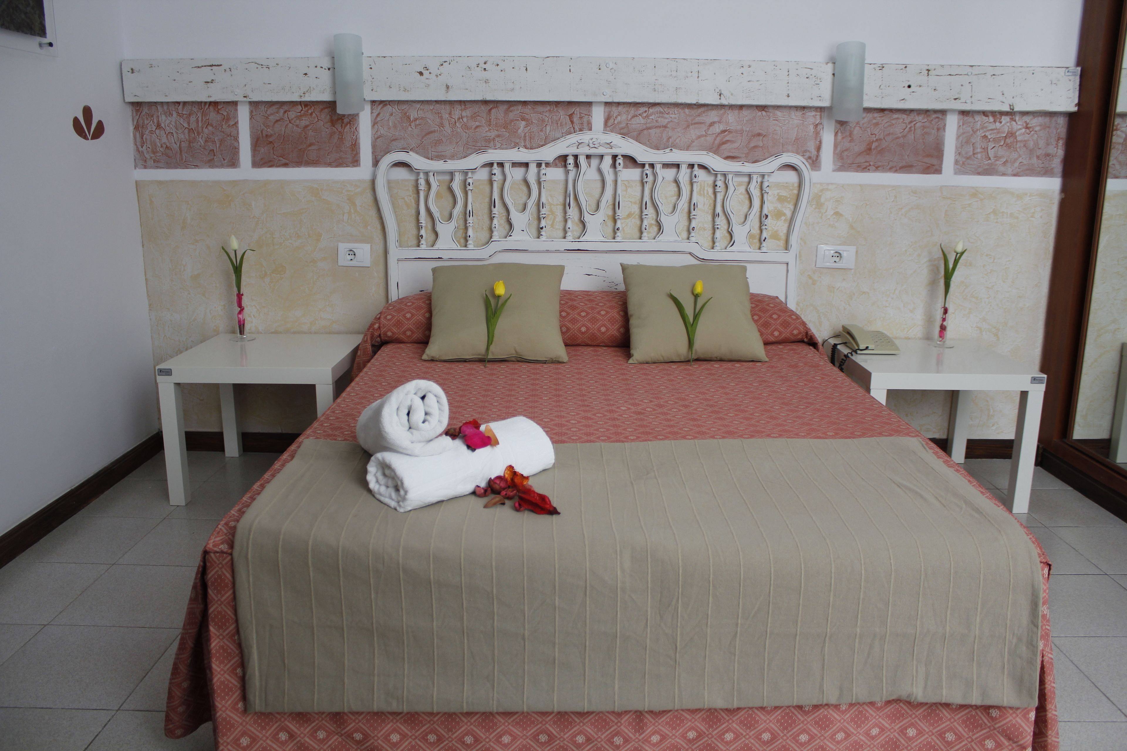 A neatly made bed with a red and beige bedspread, two pillows, a rolled towel, and a small vase with a flower on each bedside table.
