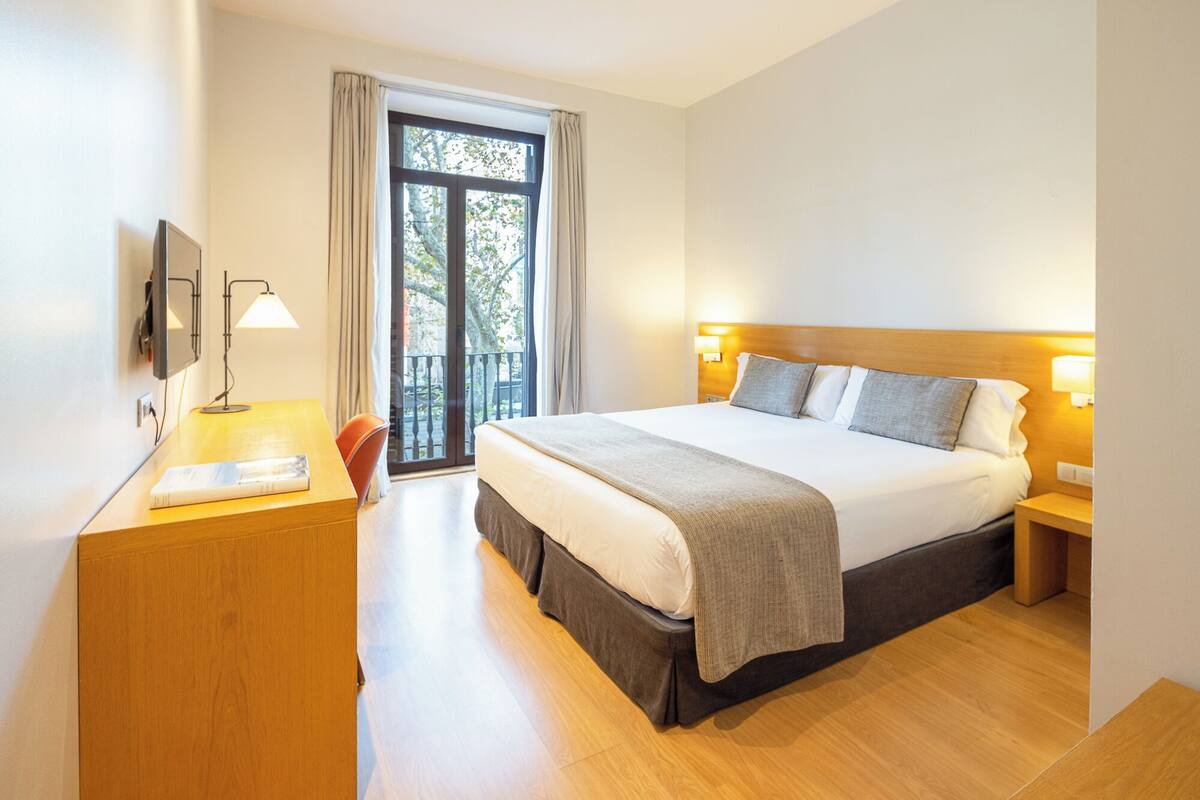Habitación de hotel con cama, lámparas de noche, un escritorio con lámpara, una silla y una ventana con cortinas.