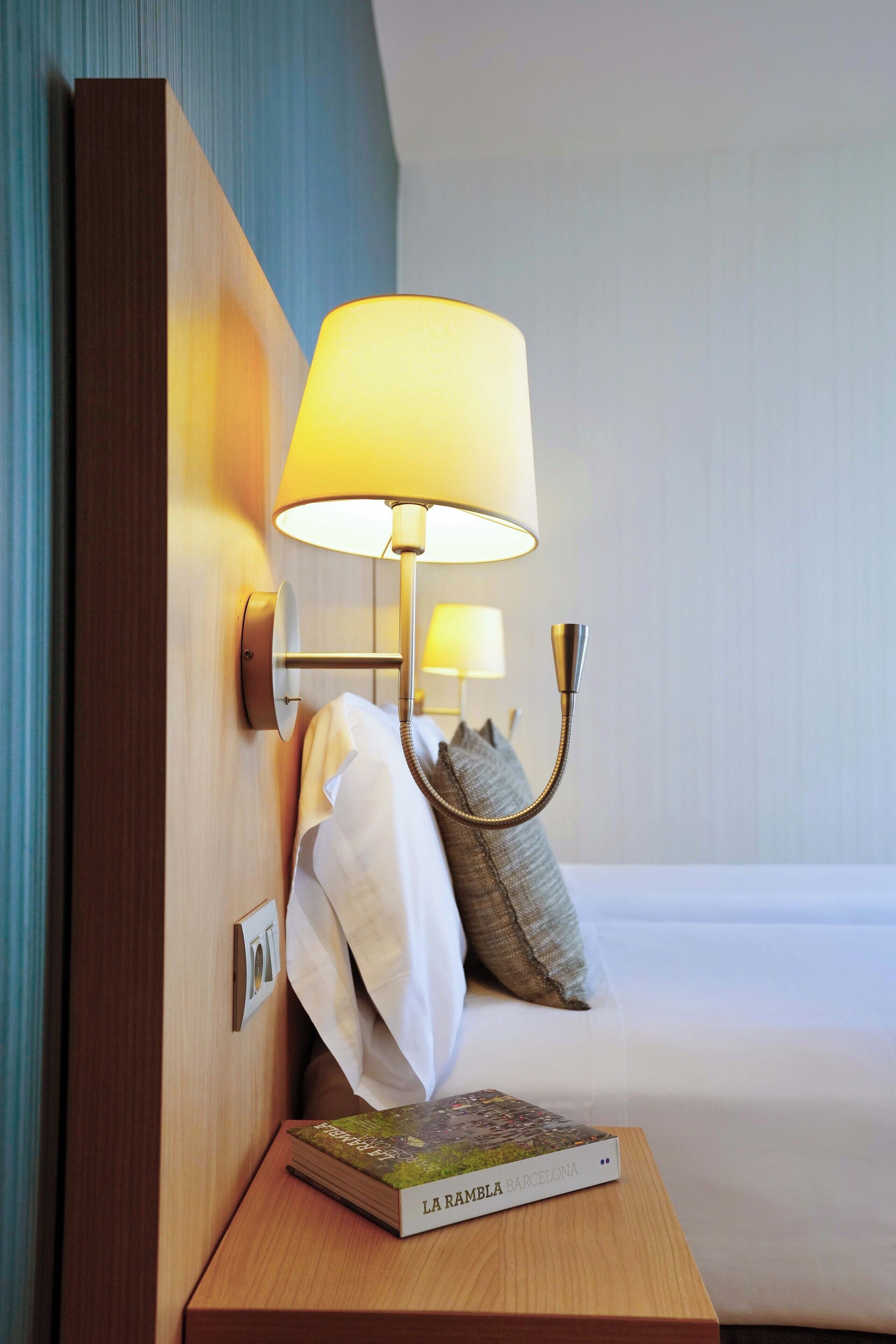 A neatly made hotel room with a bedside lamp, a book titled "La Rambla Barcelona," and a pillow on the bed.