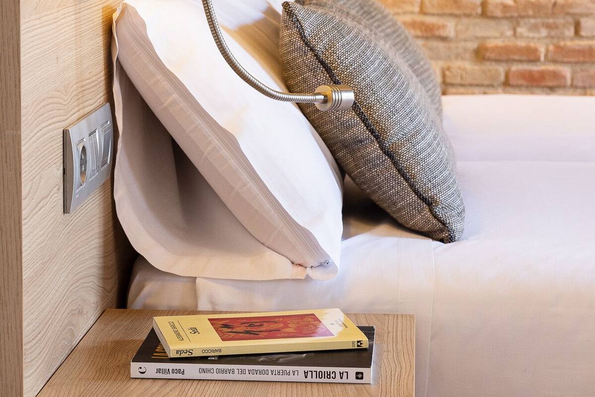 Habitación de hotel con cabecera de madera, lámpara de noche, almohada y un libro en la mesita de noche.