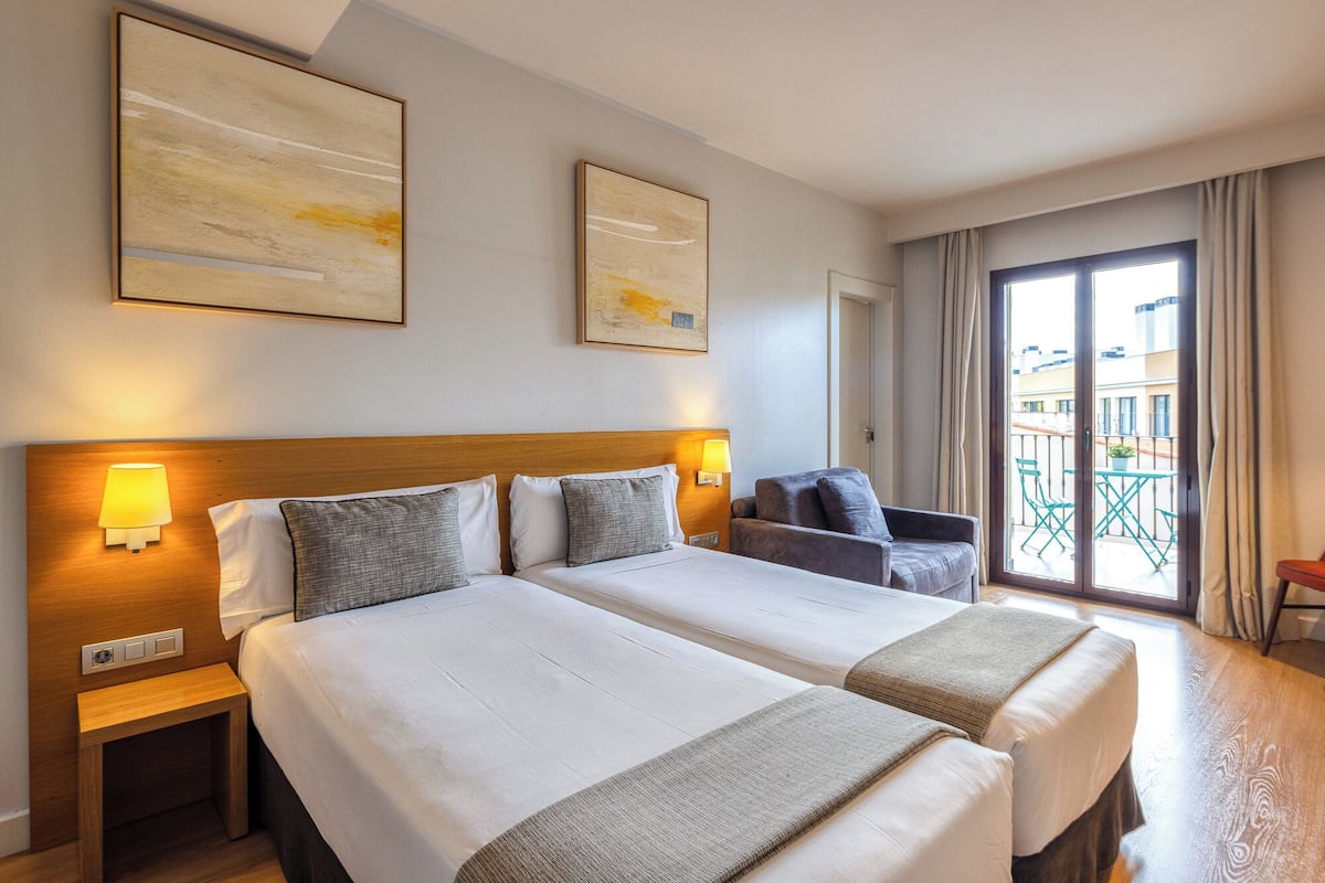 Habitación de hotel con dos camas, cabecera de madera, lámparas de noche y balcón con vistas.