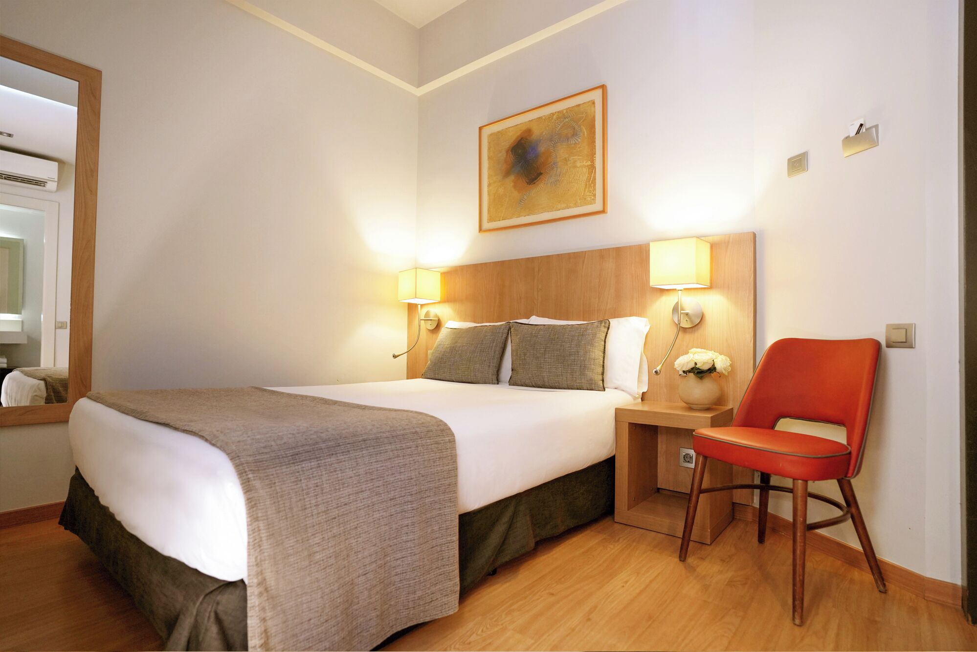 Una habitación de hotel con una cama grande, dos lámparas de noche, una mesita con un florero y una silla roja.