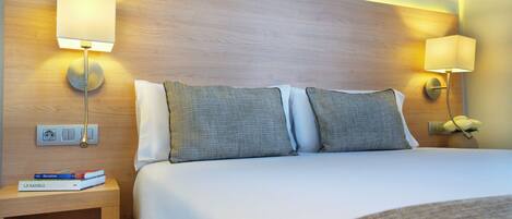 A hotel room with a bed, two bedside lamps, a nightstand with a book, and a power outlet.