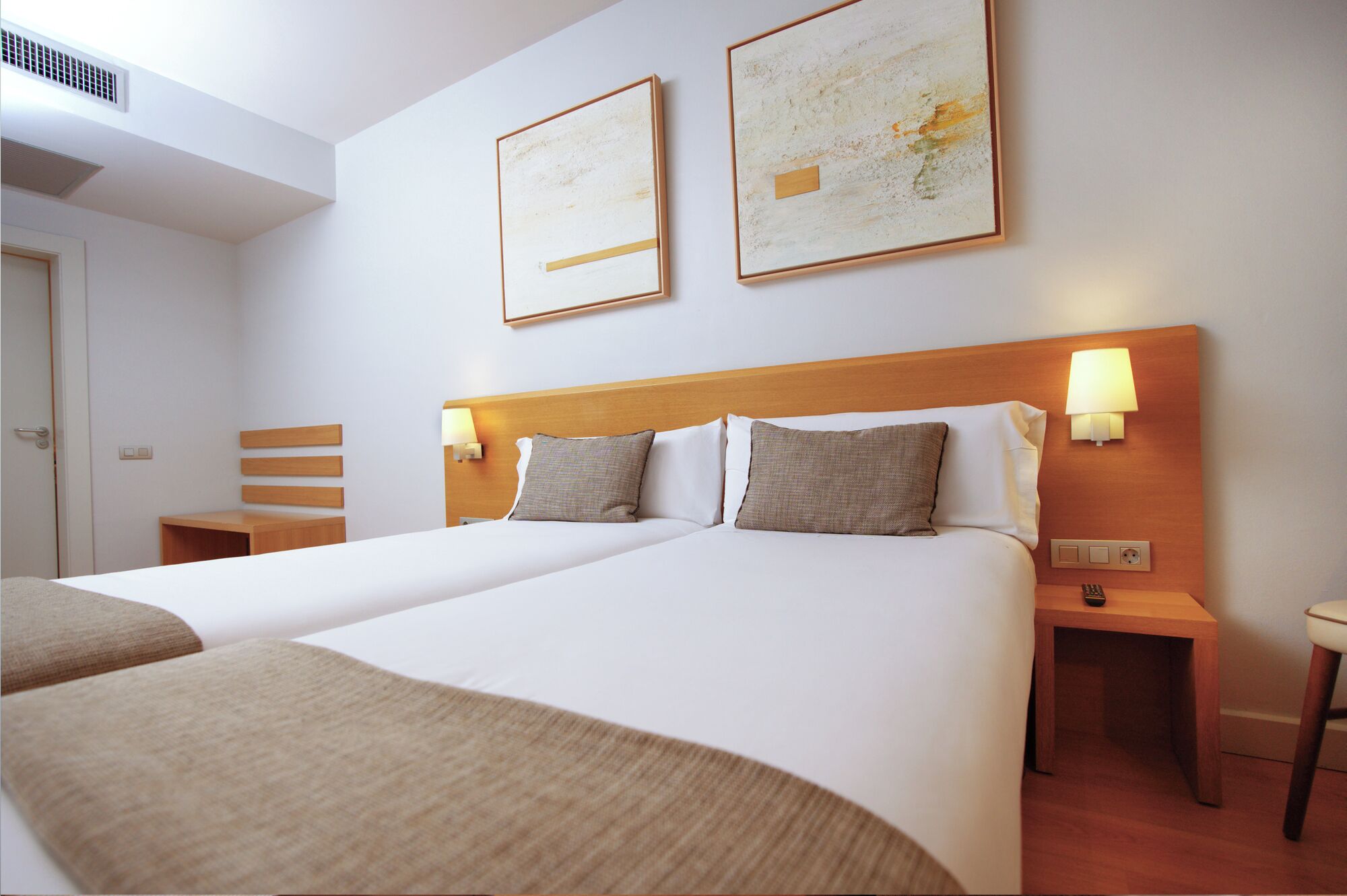 A hotel room with two beds, a wooden headboard, bedside lamps, and two framed abstract paintings on the wall.