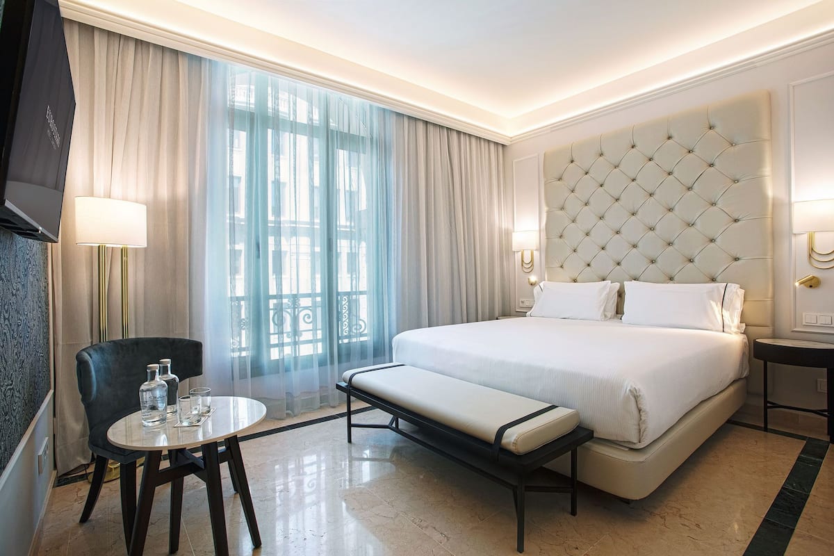 Une chambre d’hôtel avec un grand lit, une table de chevet, une chaise et une vue sur l’extérieur à travers des rideaux transparents.