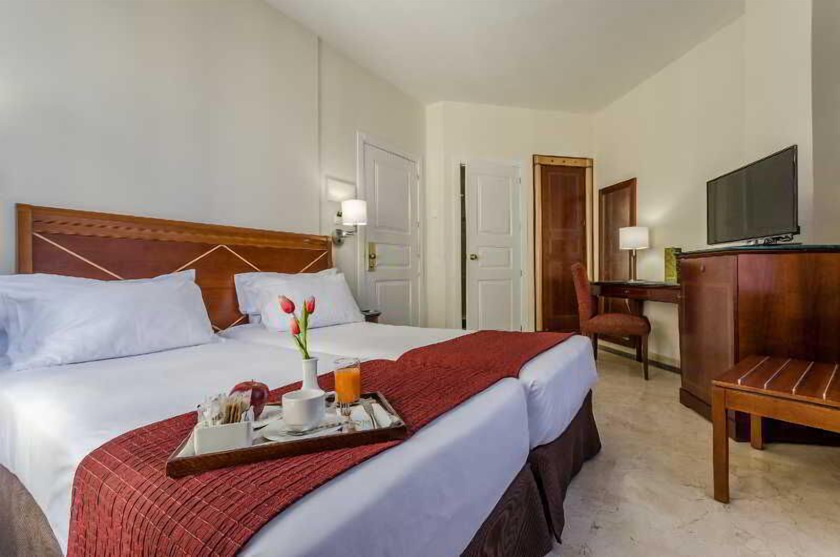 Quarto de hotel com cama, escrivaninha, cadeira, televisão e bandeja com itens de café da manhã.