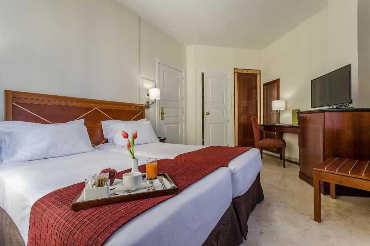 Quarto de hotel com cama, escrivaninha, cadeira, televisão e bandeja com itens de café da manhã.