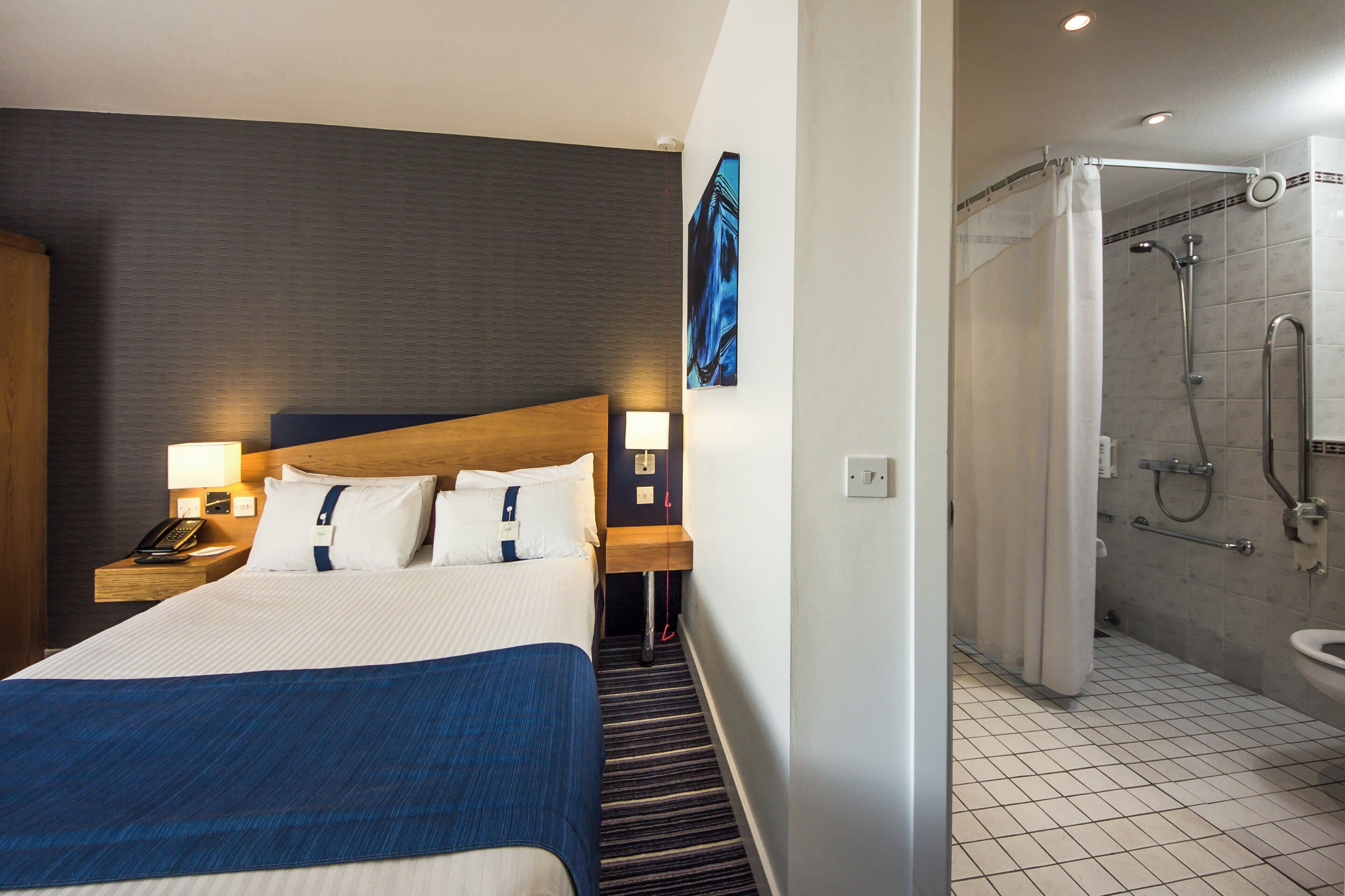 A hotel room with a bed, bedside lamps, a telephone, a painting, and a shower area.