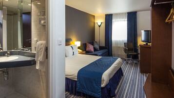 A hotel room with a large bed, a desk, a chair, a TV, a lamp, a mirror, and a bathroom with a sink and shower.