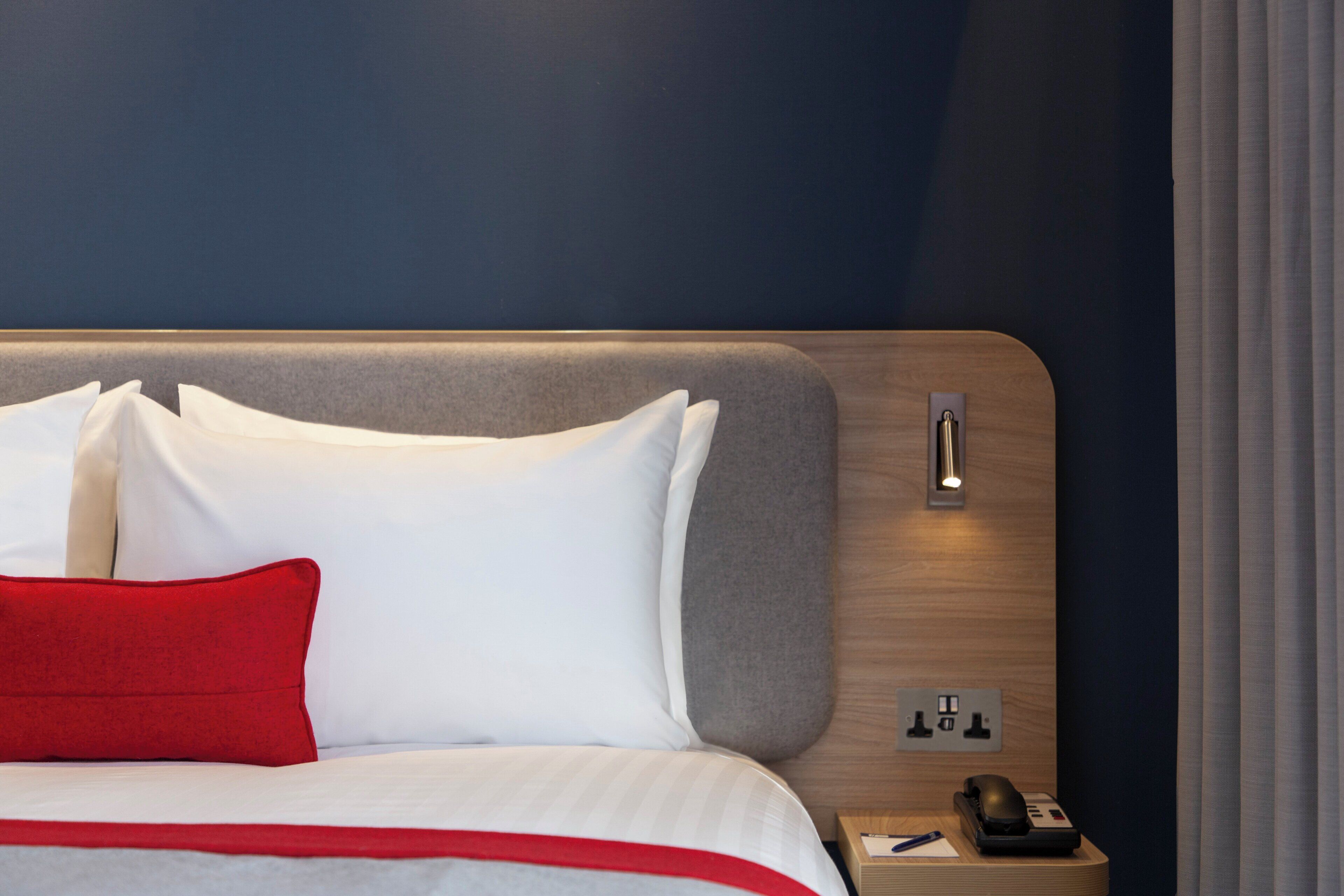 Quarto de hotel com cama, mesa de cabeceira, telefone e tomada na parede.