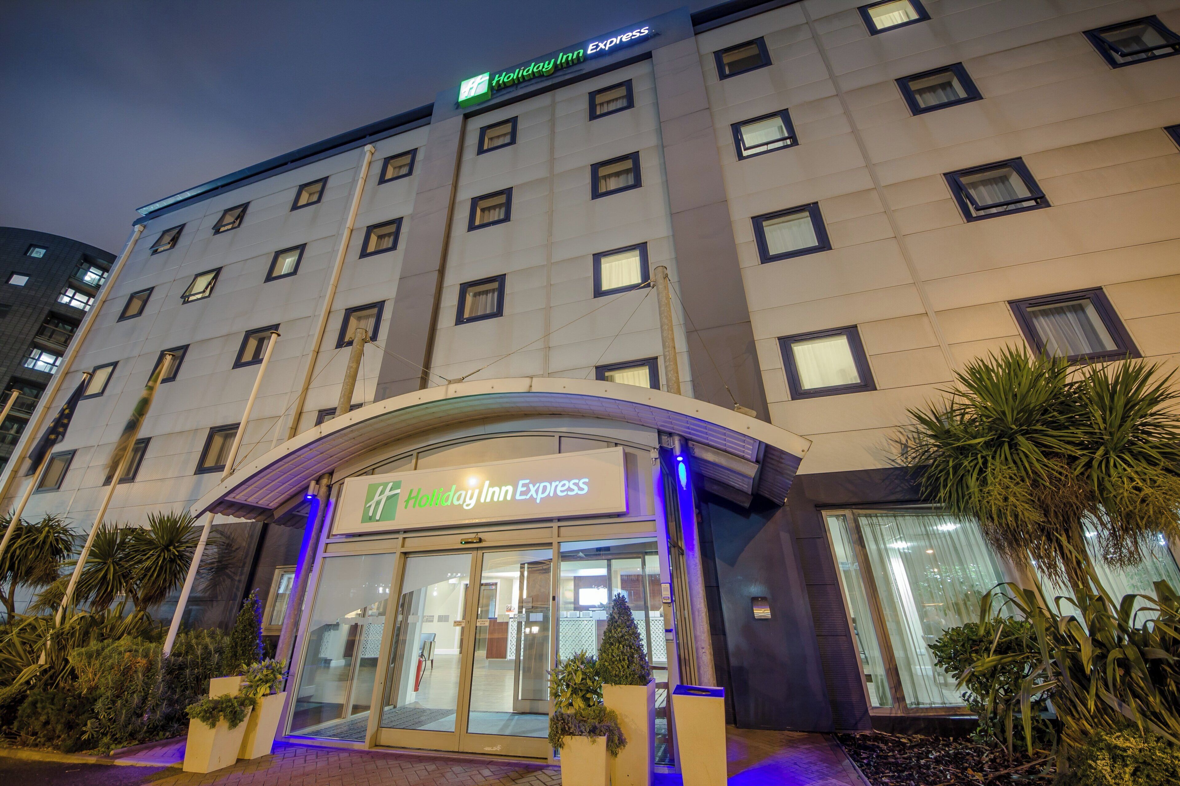L’entrée d’un hôtel Holiday Inn Express, avec une porte en verre, une enseigne au-dessus de l’entrée et une rangée de palmiers devant.