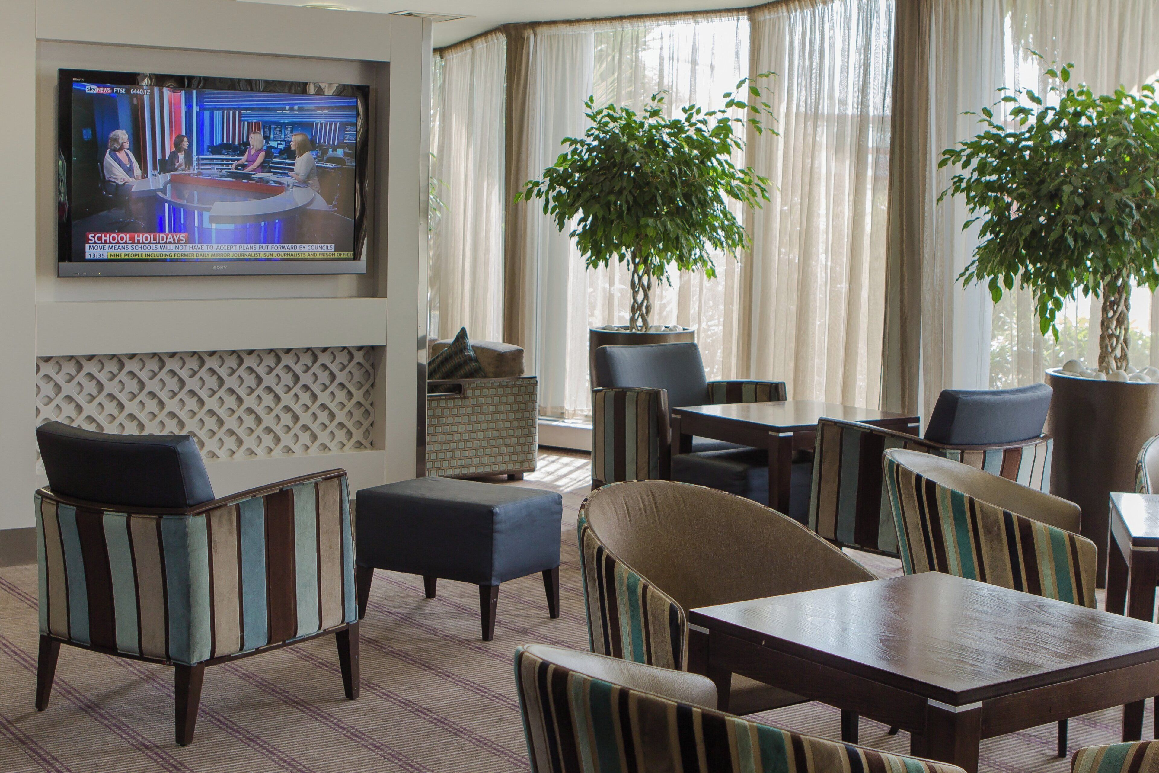 A hotel room with a television displaying a news program, a seating area with striped chairs and ottomans, and large windows with sheer curtains.