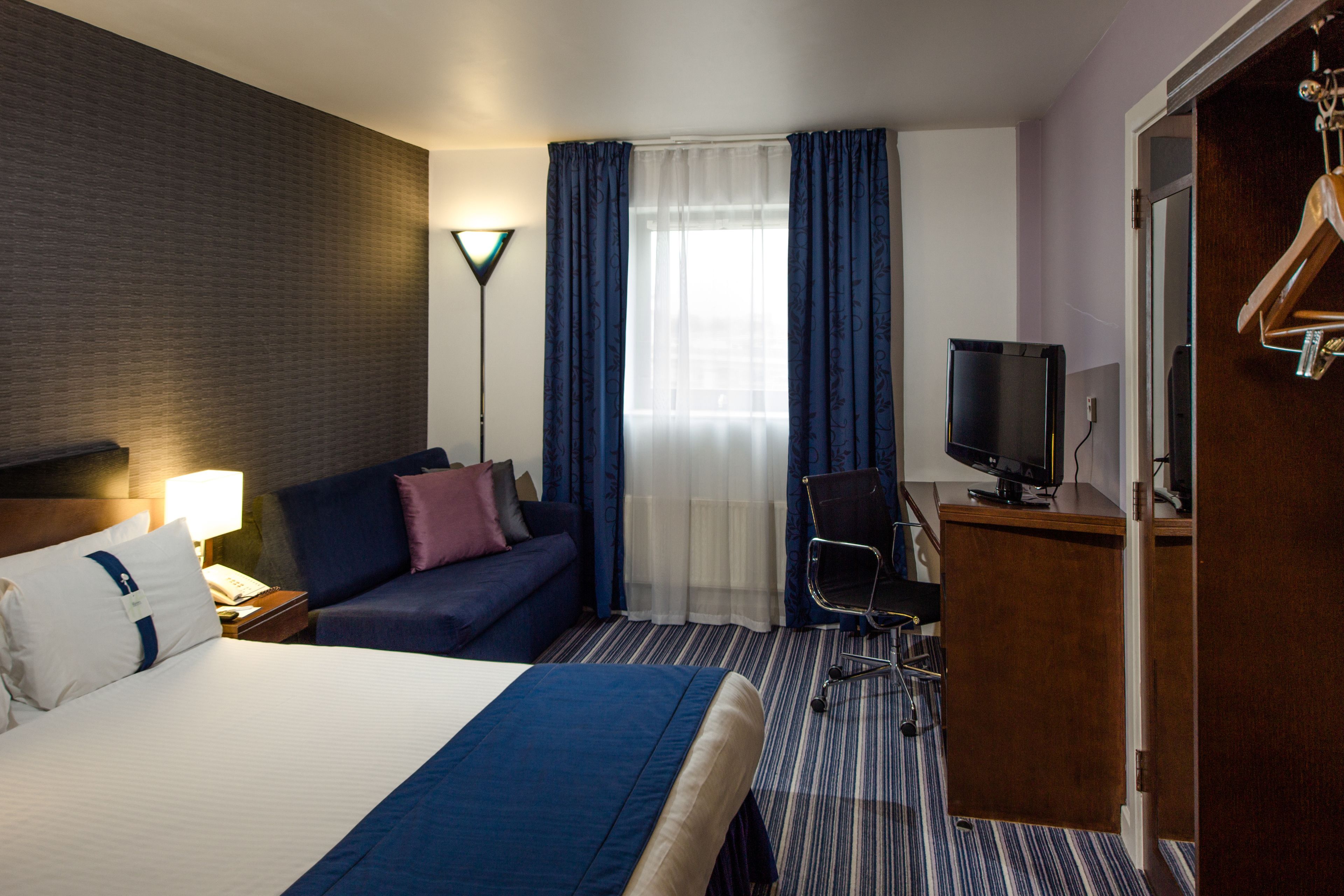 A hotel room with a large bed, a sofa, a desk with a television, and a wardrobe.